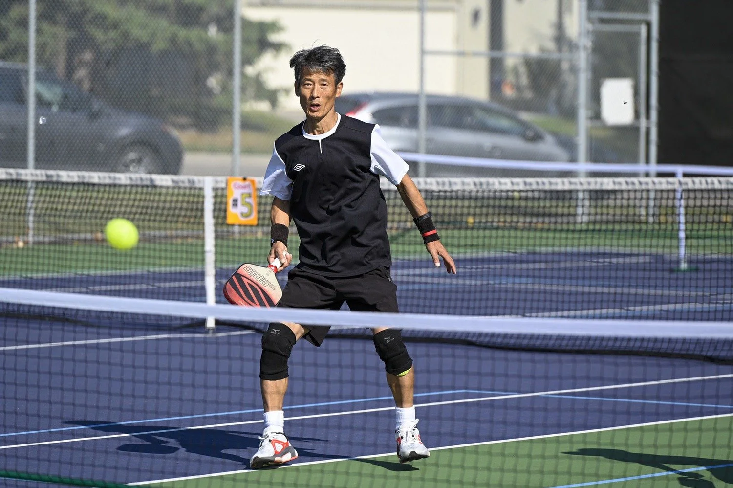 A person with athletic gear playing pickleball on an outdoor court, holding a paddle, with a yellow ball in play.