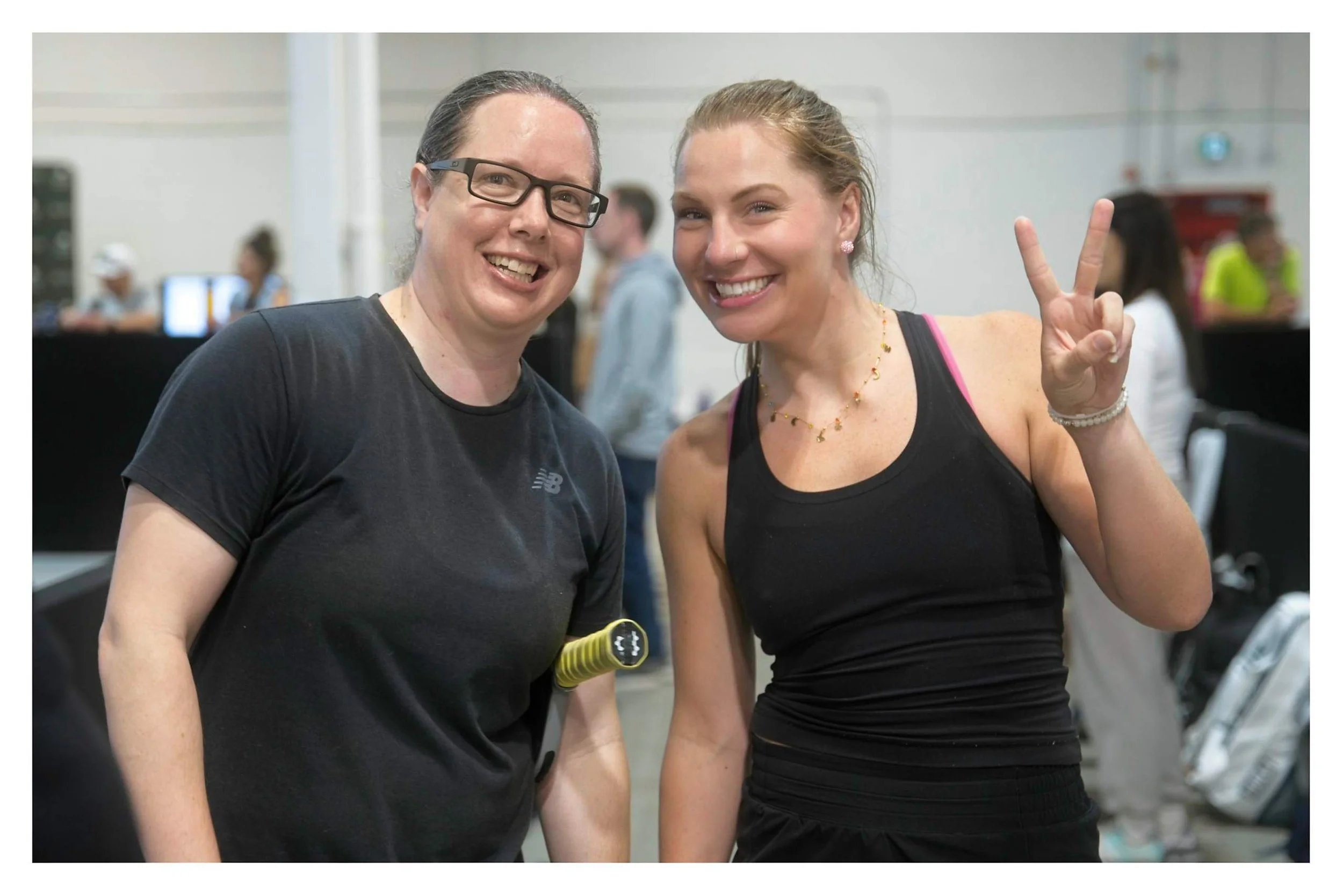 Two women smiling at the camera, one showing a peace sign, in a gym or fitness setting with other people in the background.