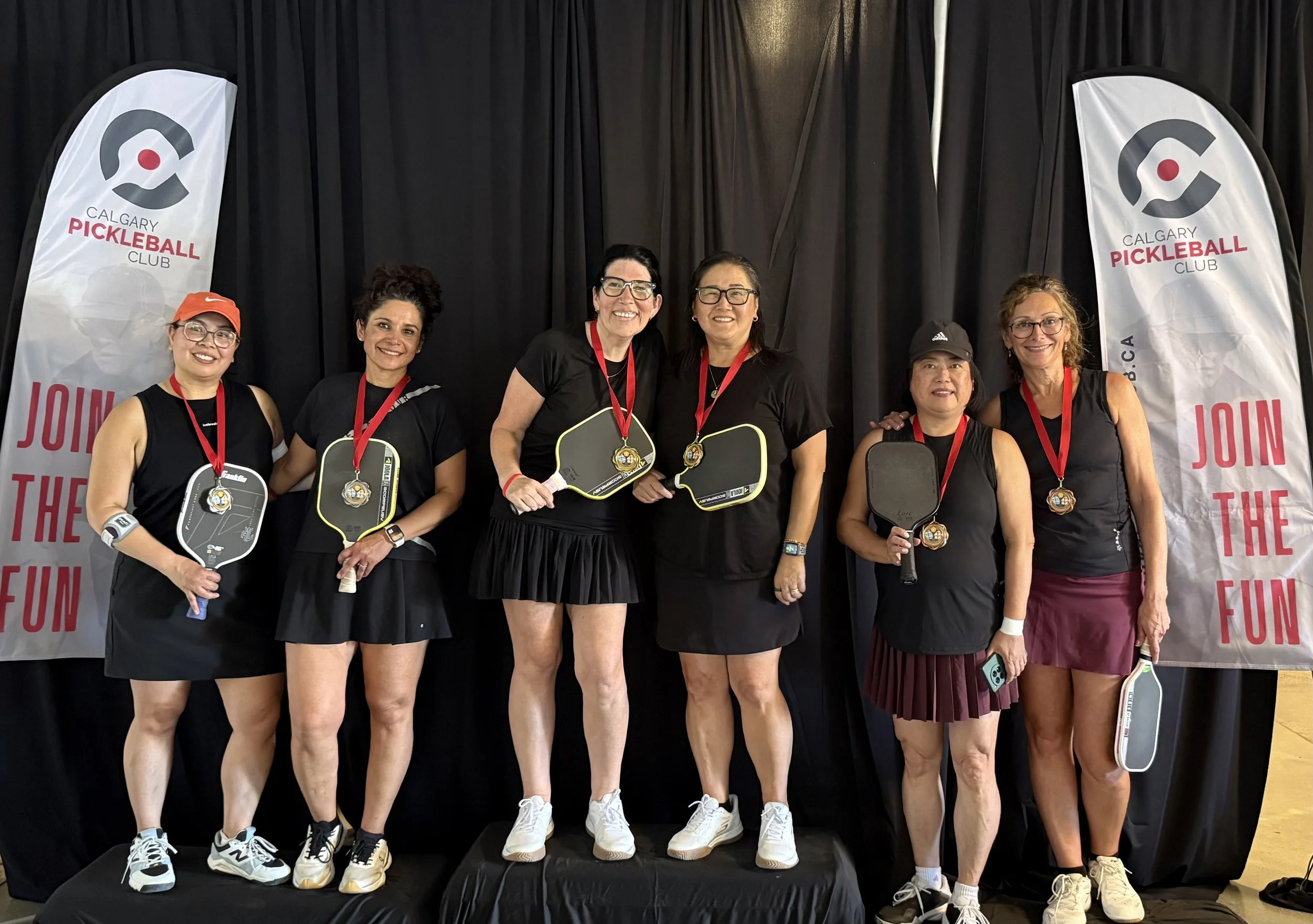 A group of seven women standing together on a platform, holding pickleball paddles, and wearing medals around their necks. They are dressed in athletic clothing, and the background features banners for the Calgary Pickleball Club.