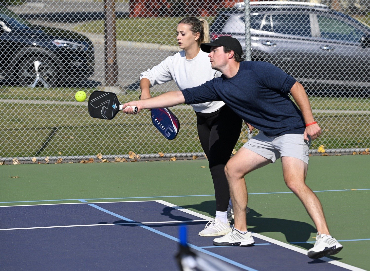 A man and a woman playing pickleball on an outdoor court. The man is hitting the ball with a paddle while the woman is watching. The court is enclosed by a chain-link fence, with parked cars and grass in the background.