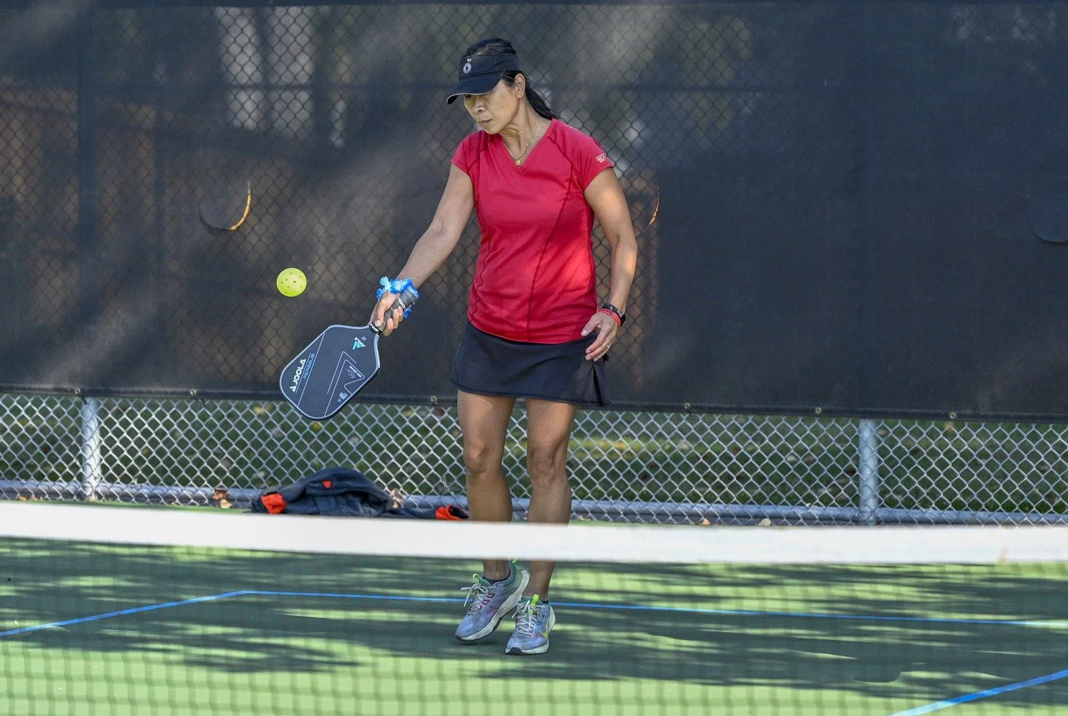 A woman on a tennis court preparing to hit a tennis ball with a paddle, wearing a red sports shirt, black skirt, and gray sneakers.