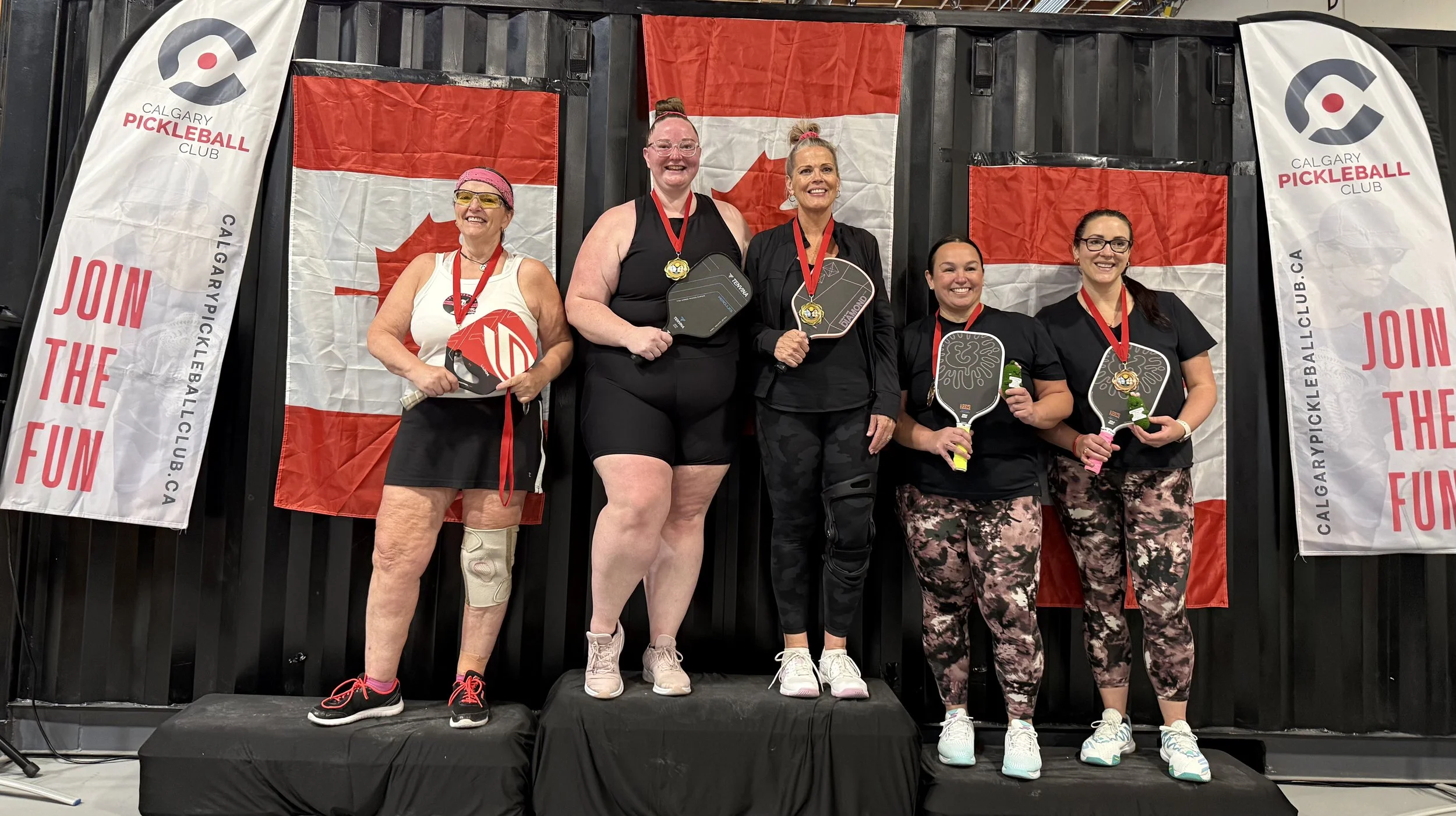Group of five women standing on a winners' podium holding pickleball paddles, with Canadian flags and banners in the background, celebrating their victories at a pickleball event.