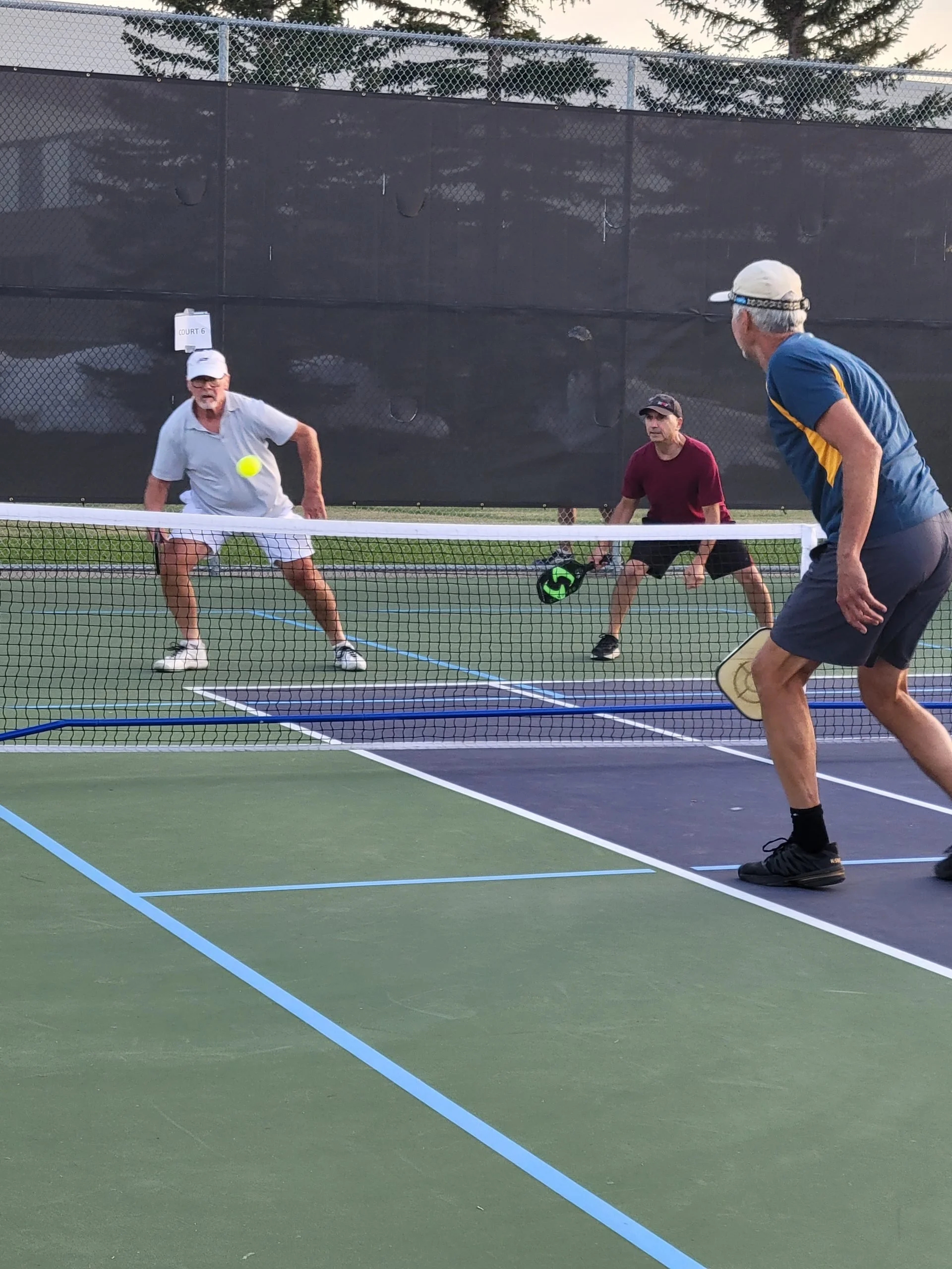 Three men playing pickleball on a court, one preparing to hit the ball while the other two watch, with a black windscreen on the fence behind them.