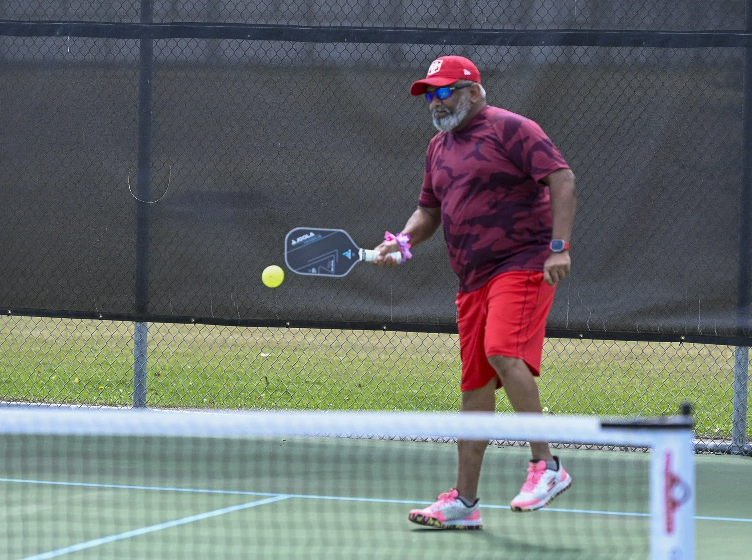 A man playing pickleball on a court with a black fence in the background. He is wearing a red cap, sunglasses, a camouflage-patterned red shirt, red shorts, and pink athletic shoes, holding a pickleball paddle and preparing to hit a yellow pickleball