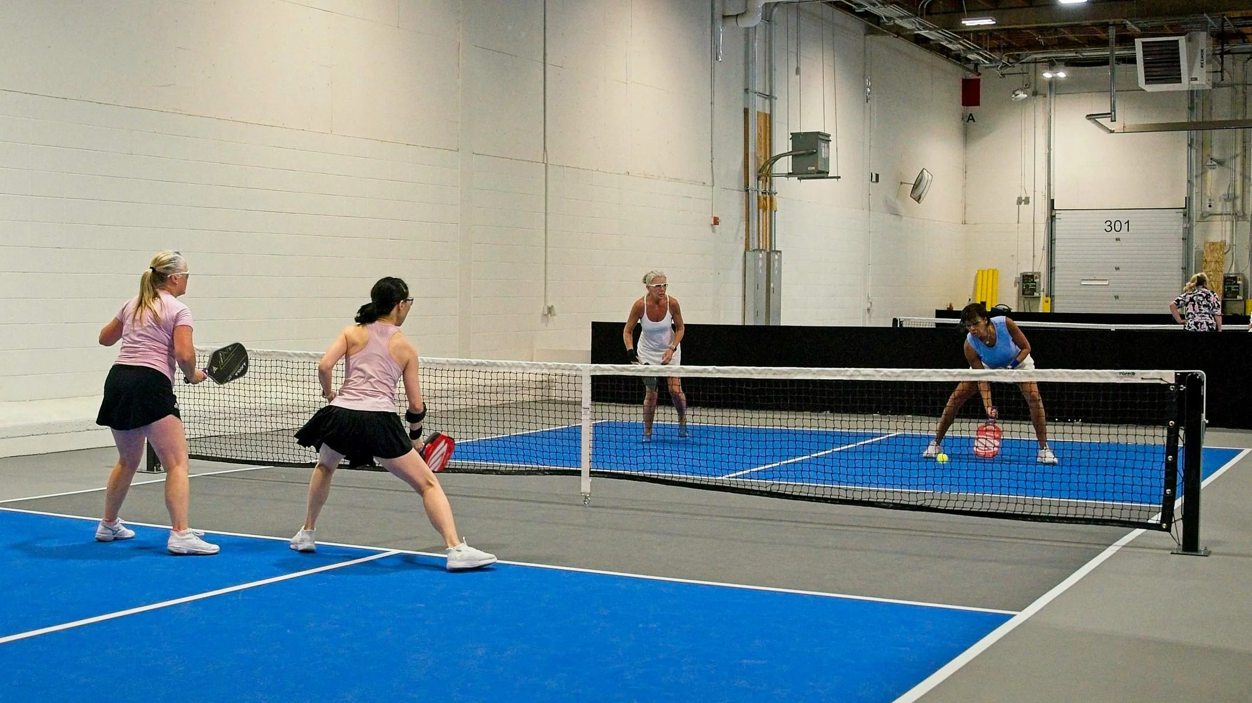 Women playing pickleball indoors on a blue court, with a net, engaged in a game, some spectators in background.