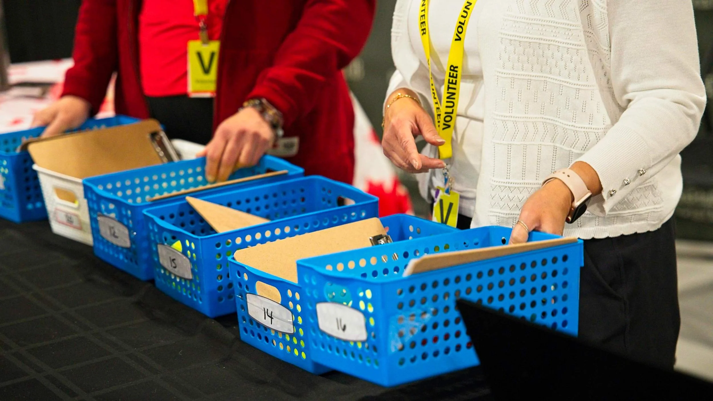 People organizing blue plastic baskets with files and clipboards on a black table, wearing volunteer badges around their necks.