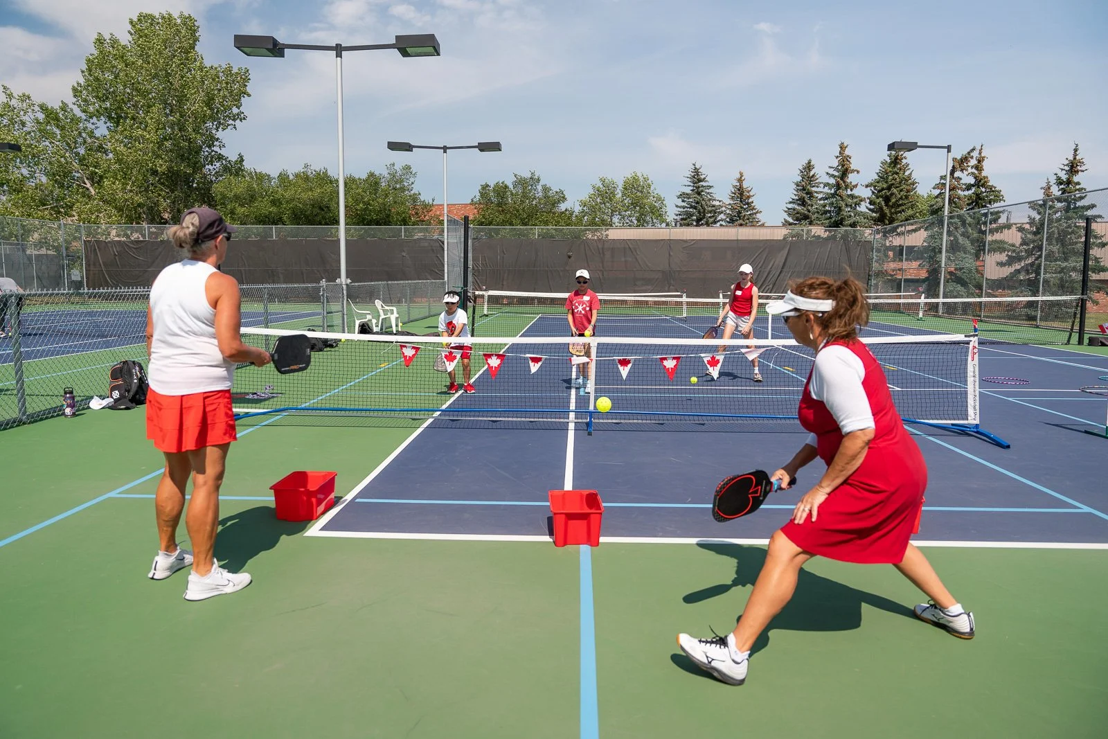 Two women and three children playing pickleball on outdoor courts with a net, tennis balls, and paddles on a sunny day.