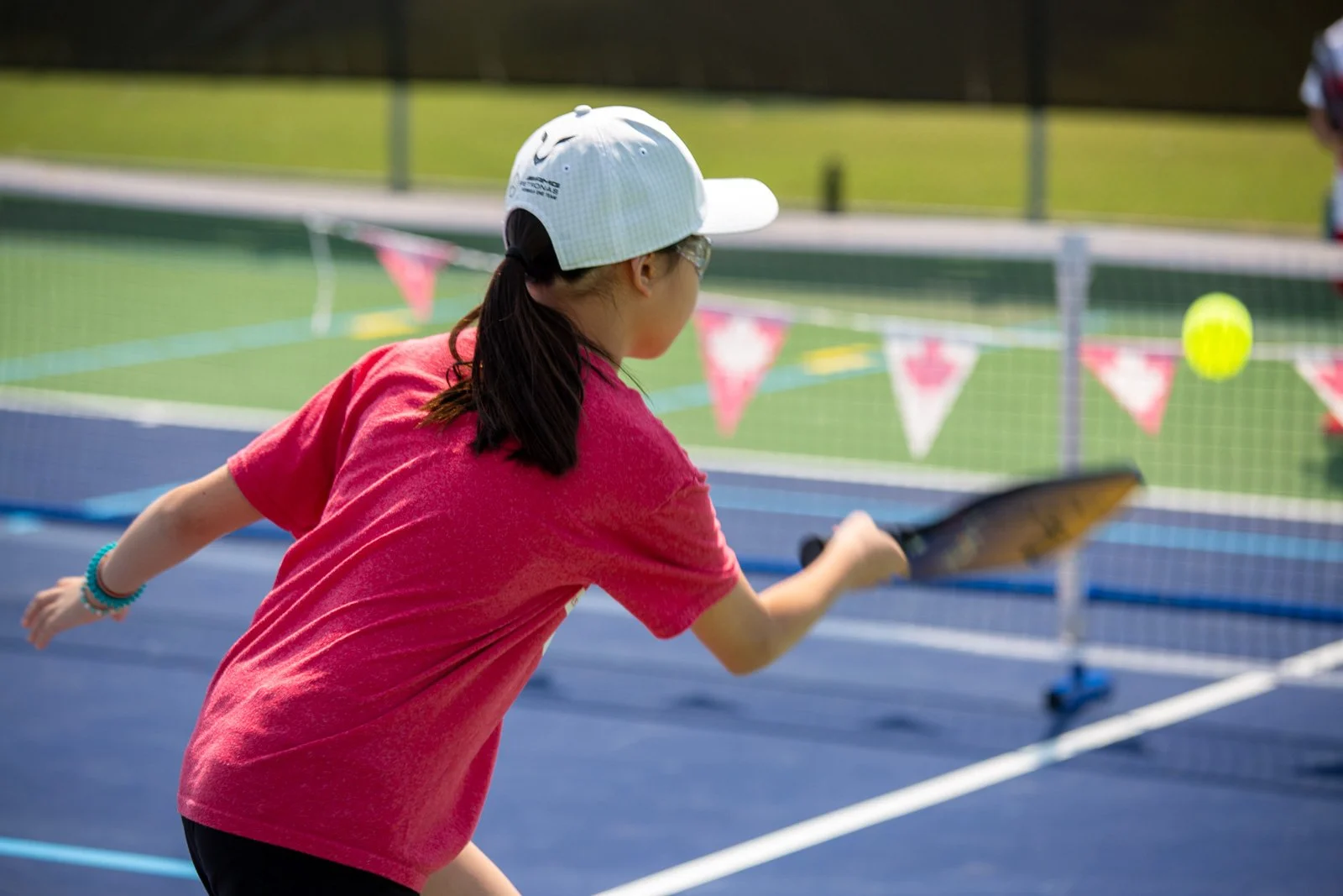 Child playing tennis on an outdoor court, wearing a pink shirt and a white cap, hitting a yellow tennis ball.
