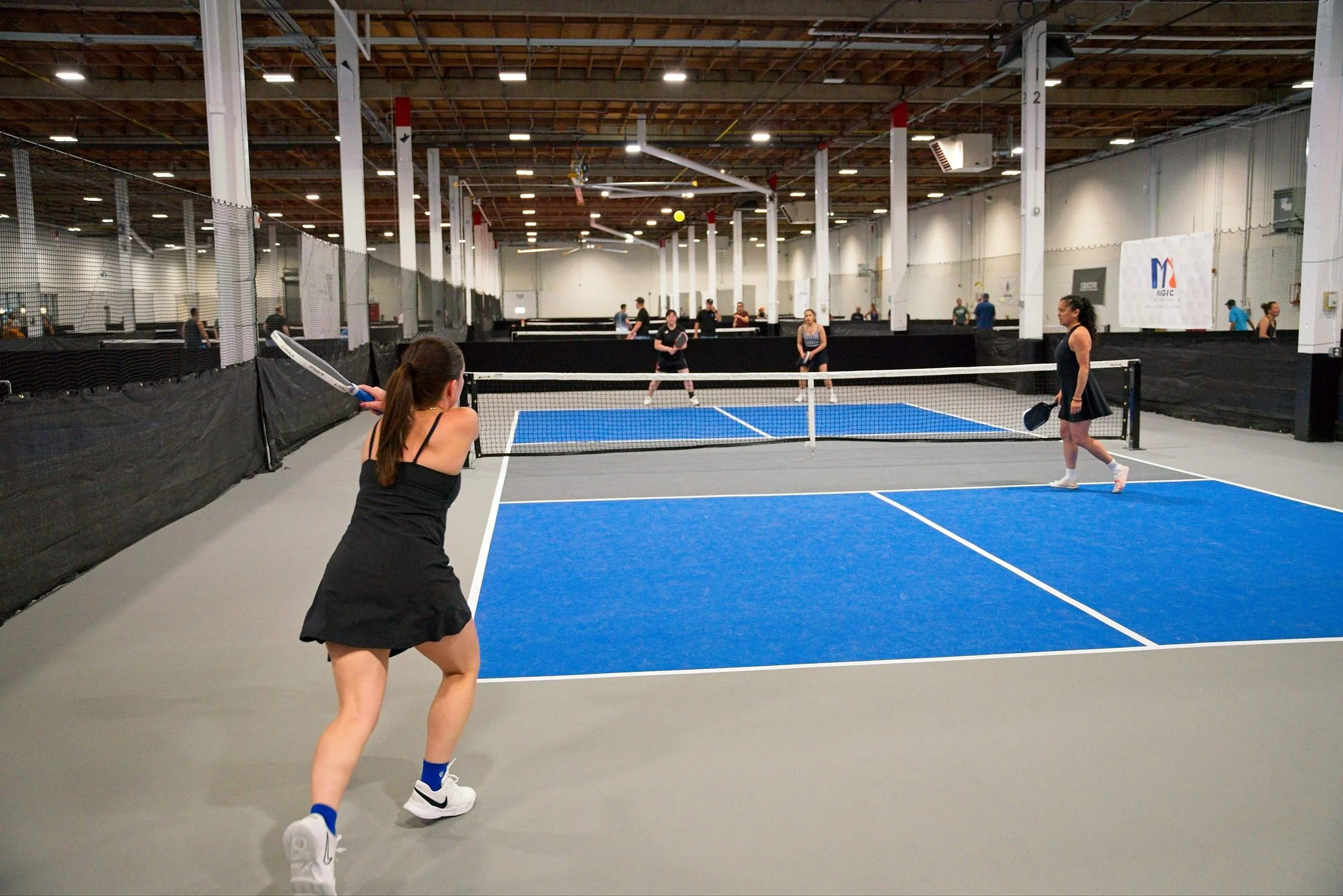 People playing pickleball on indoor courts, with some preparing to hit the ball and others waiting, under a high ceiling with exposed wood beams.