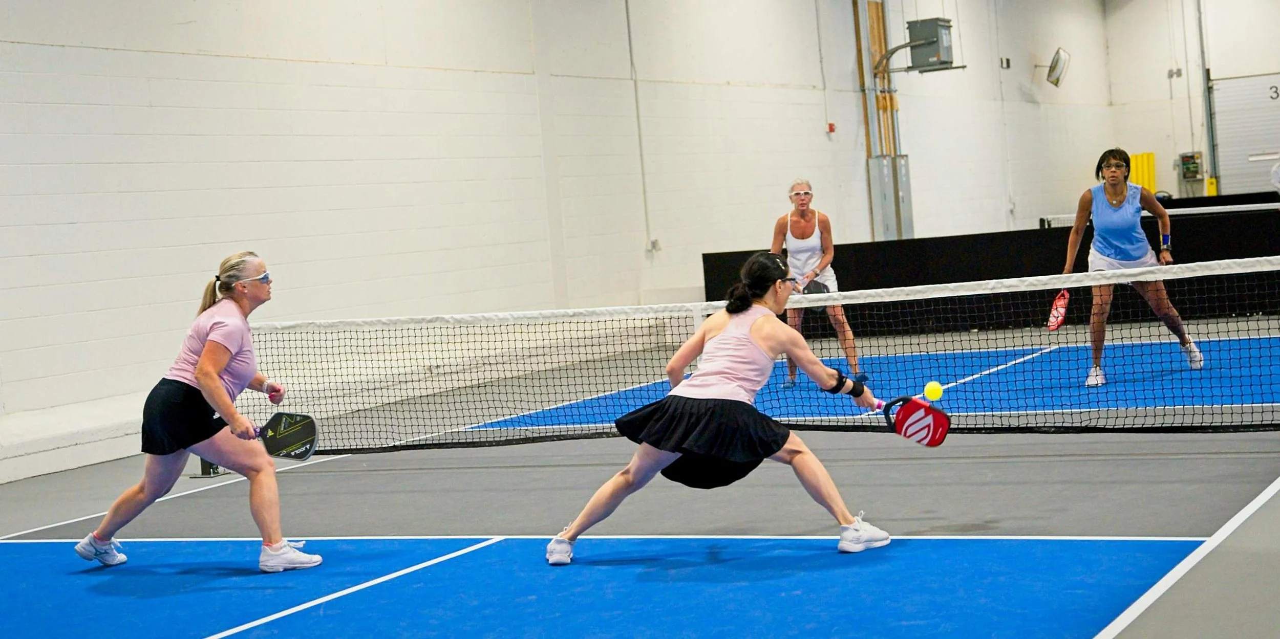 Four women playing pickleball on an indoor court divided by a net, with two women in pink shirts and black skirts on the near side, and two women in white and blue on the far side, one of whom is ready to return a shot.