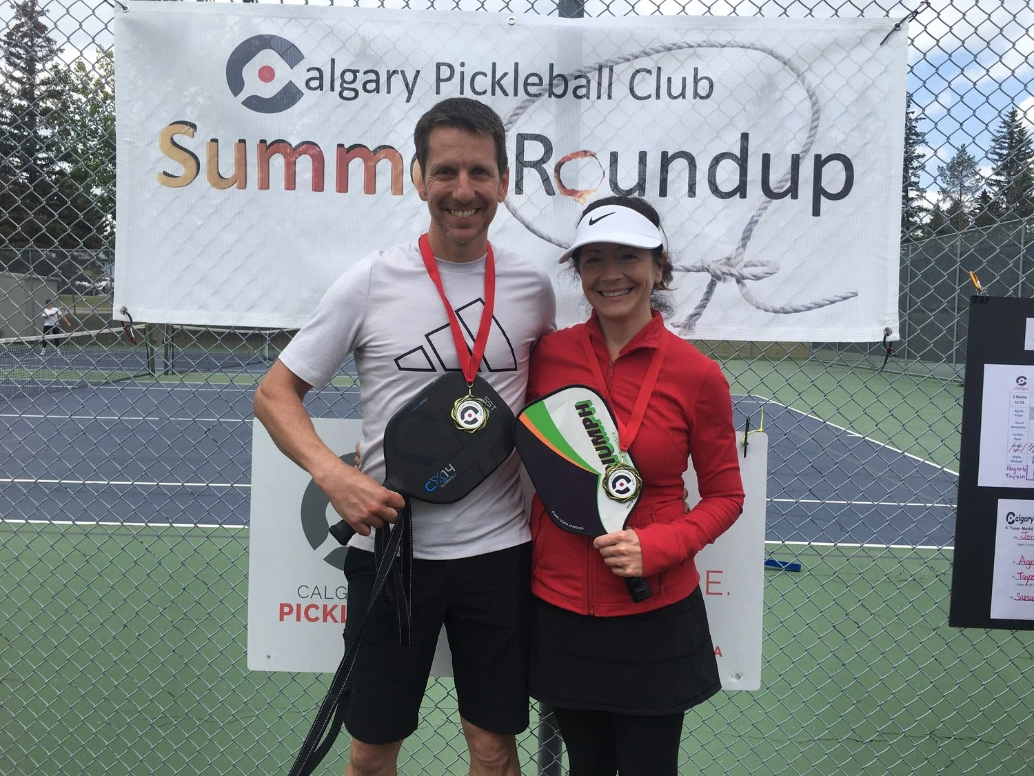 Two people, a man and a woman, standing on a tennis court in front of a banner that reads 'Calgary Pickleball Club Summer Roundup.' They are smiling and wearing medals around their necks, holding pickleball paddles.