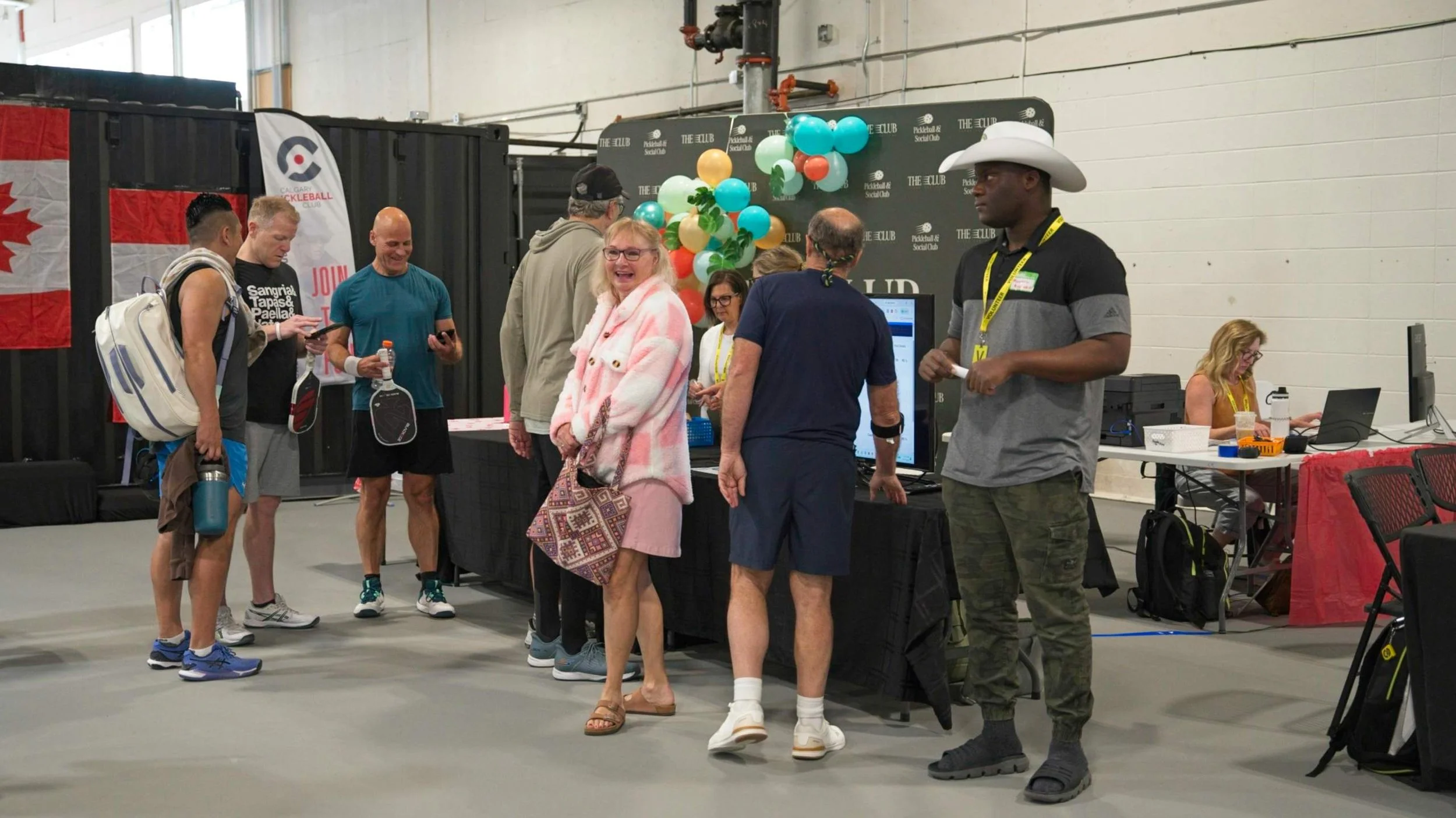 Group of people at an indoor event, standing near a booth with colorful balloons and a digital screen, with a woman wearing a pink coat and glasses smiling at the camera.