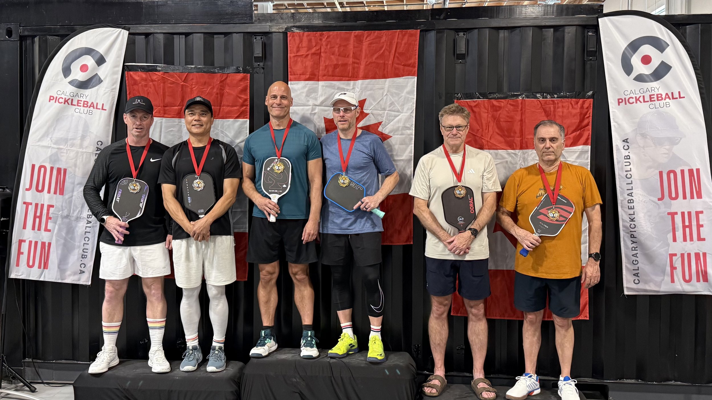 Six men standing on a podium with medals around their necks, holding pickleball paddles. Behind them are Canadian flags and banners for Calgary Pickleball Club. The men are dressed in athletic wear, and they are smiling at the camera.