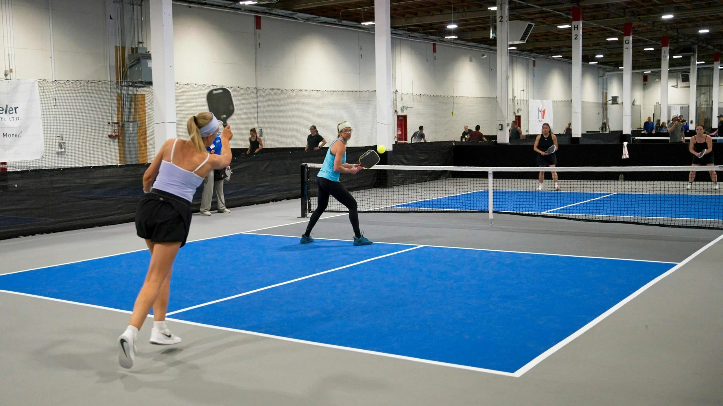 Women playing pickleball on an indoor court with multiple games happening in the background.