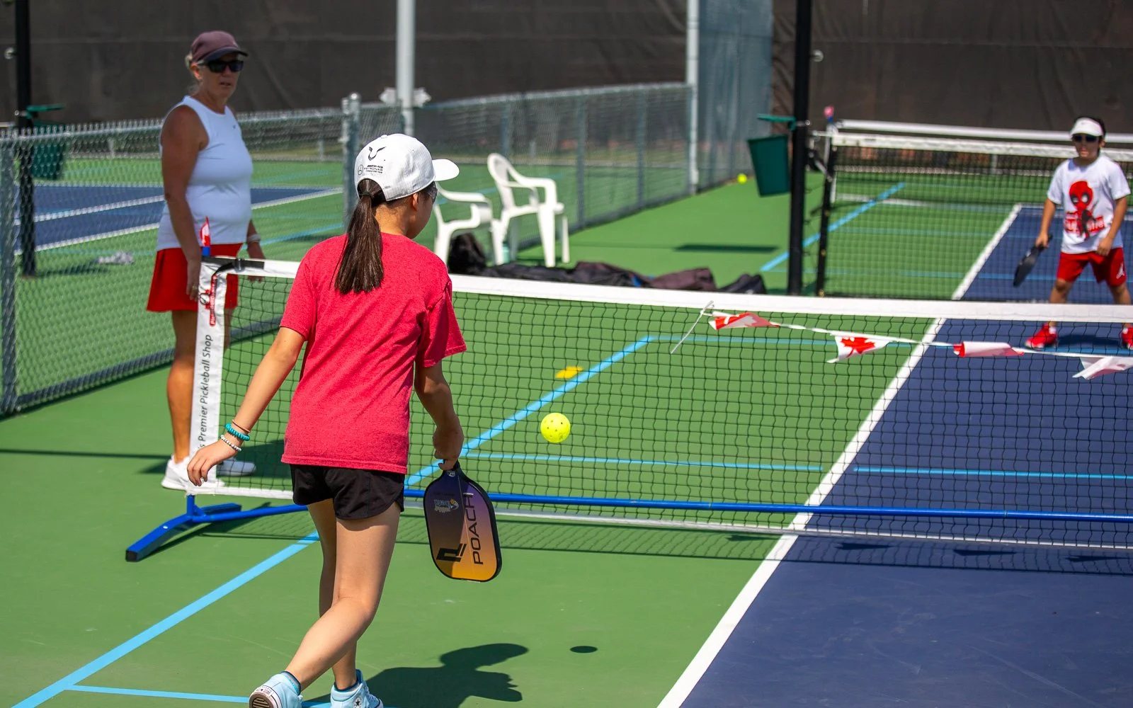 Children playing pickleball on outdoor courts, with a woman standing nearby, holding pickleball paddles, and a net with small flags hanging.