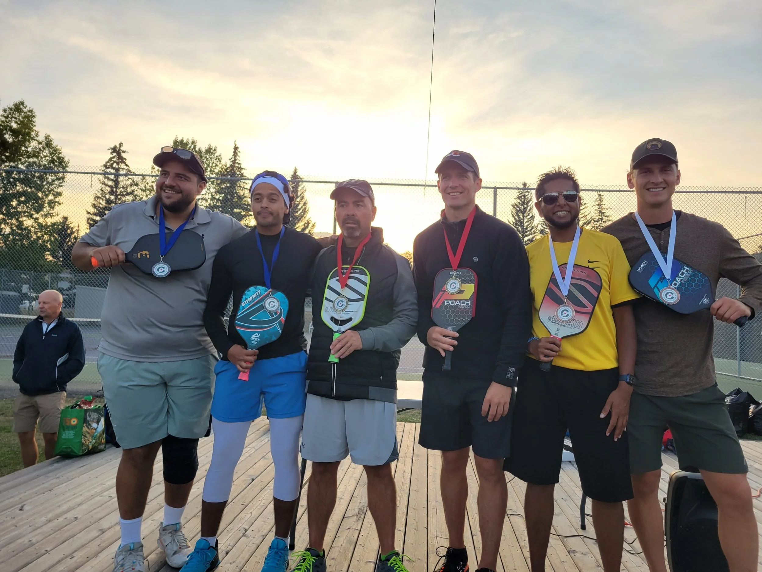 Group of six men on a tennis court with medals around their necks, standing side by side after a match, with a sunset and trees in the background.