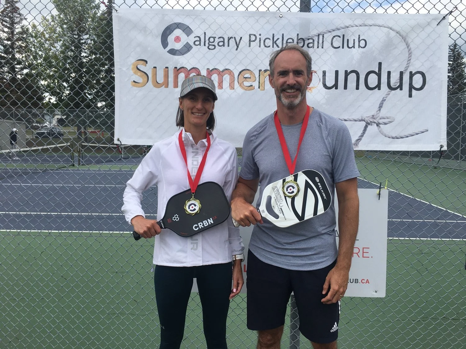 Two people standing in front of a fence with a banner that reads 'Calgary Pickleball Club Summer Roundup.' They are holding pickleball paddles, wearing medals, and smiling. The woman is dressed in a white jacket and cap, while the man wears a gray t-