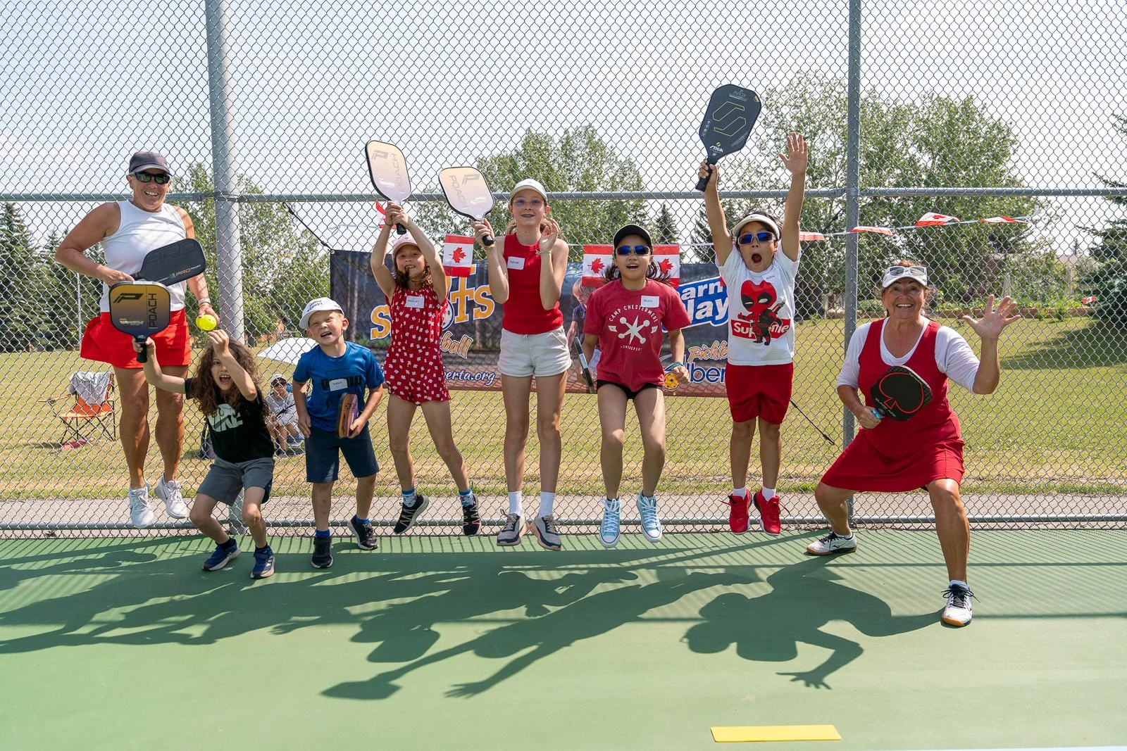 Group of children and adults celebrating on a tennis court, holding paddles and tennis balls, with a chain-link fence and banners in the background.