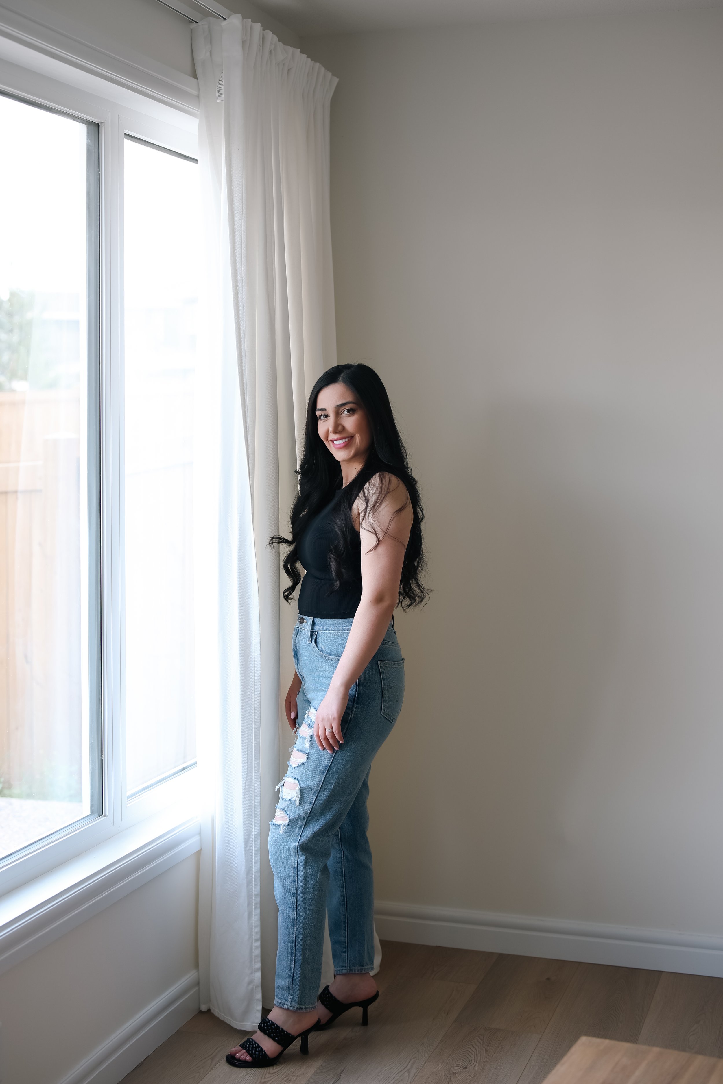 A woman with long dark hair, wearing a black sleeveless top, ripped jeans, and black high heels, standing indoors next to a window with white curtains in a room with light-colored walls and wooden flooring.
