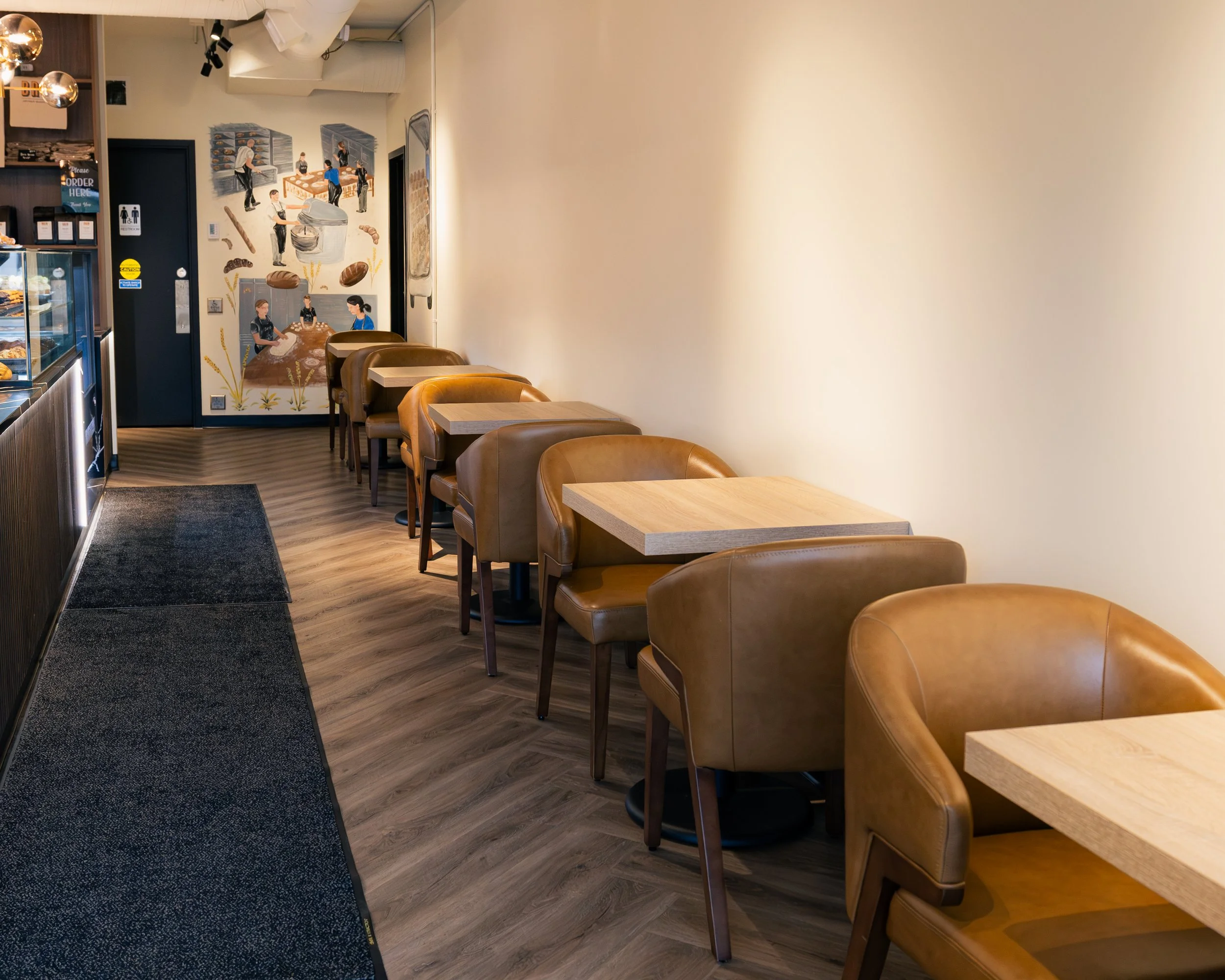 Empty chairs and tables in a cozy cafe with wall art depicting bakery scenes.