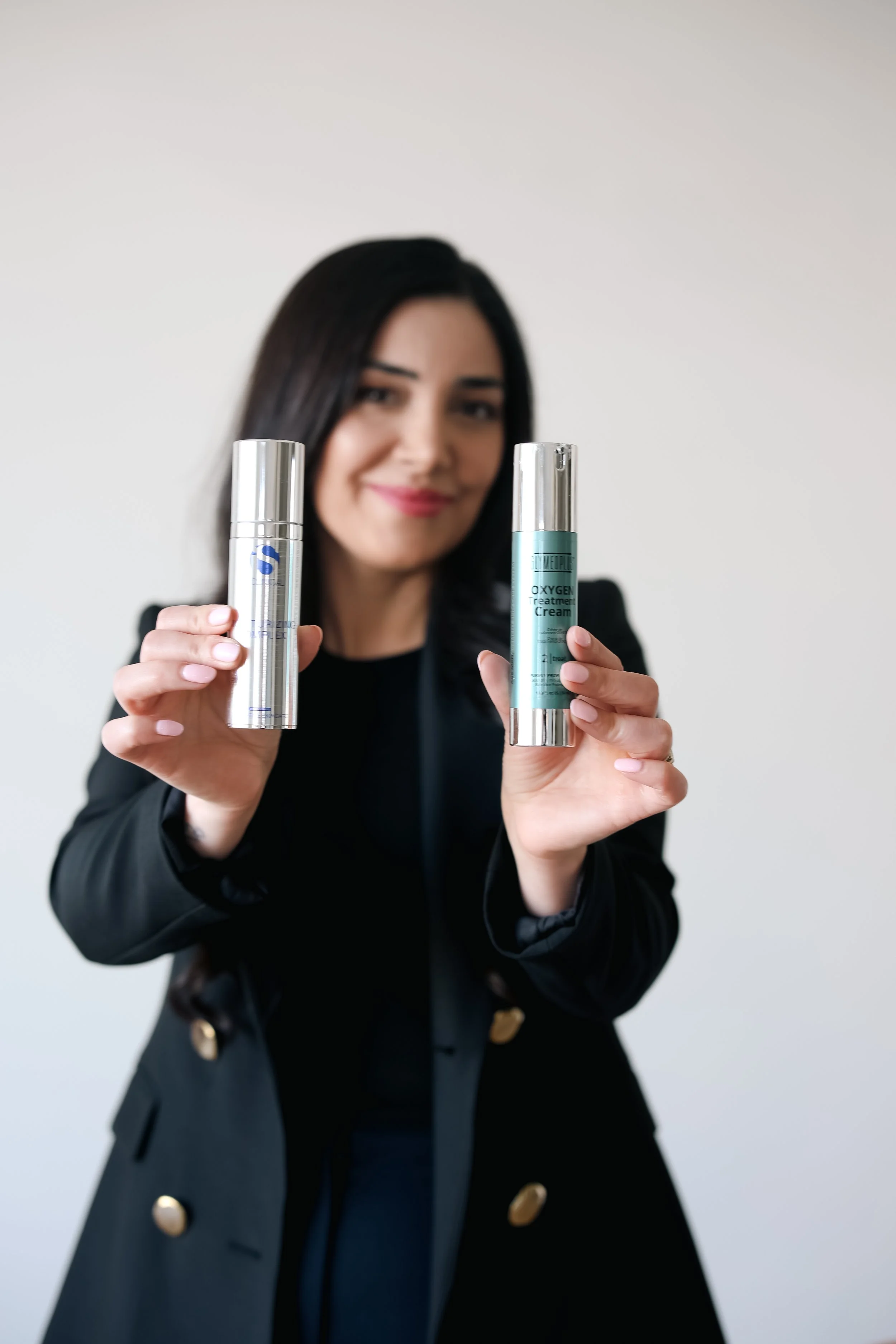 A woman with dark hair wearing a black blazer holding skincare products in her hands, smiling at the camera.