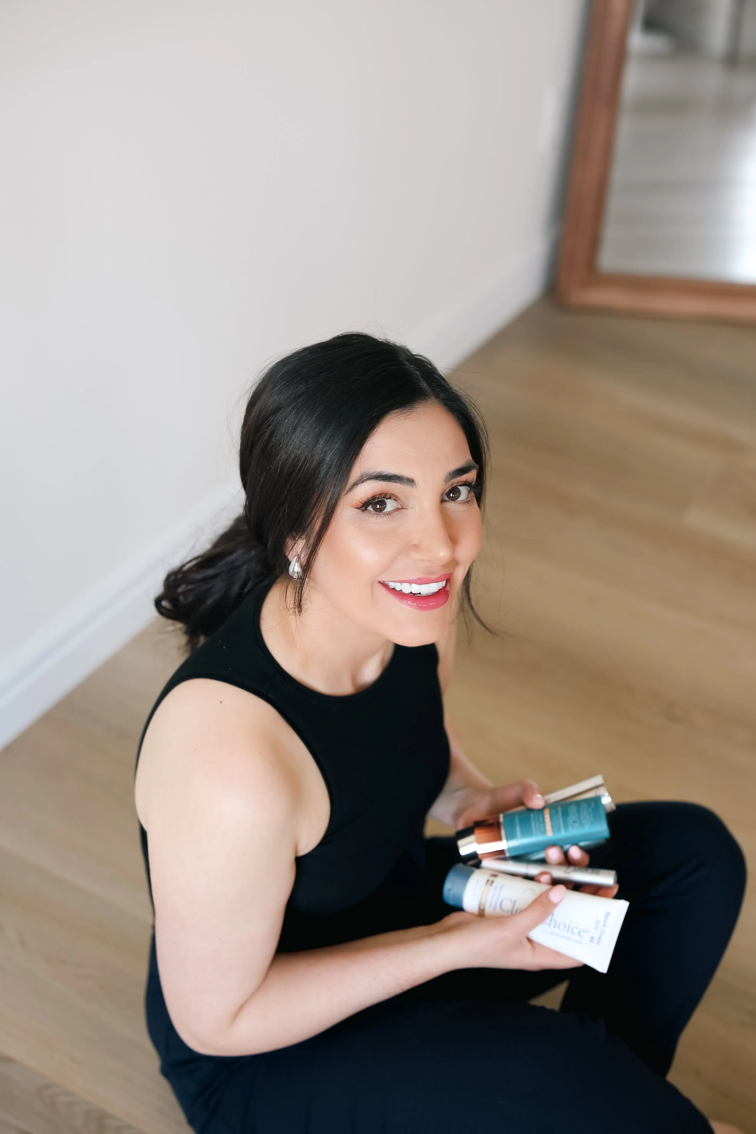 A woman with dark hair wearing a black sleeveless top, sitting on a wooden floor, smiling and holding several skincare or beauty products.