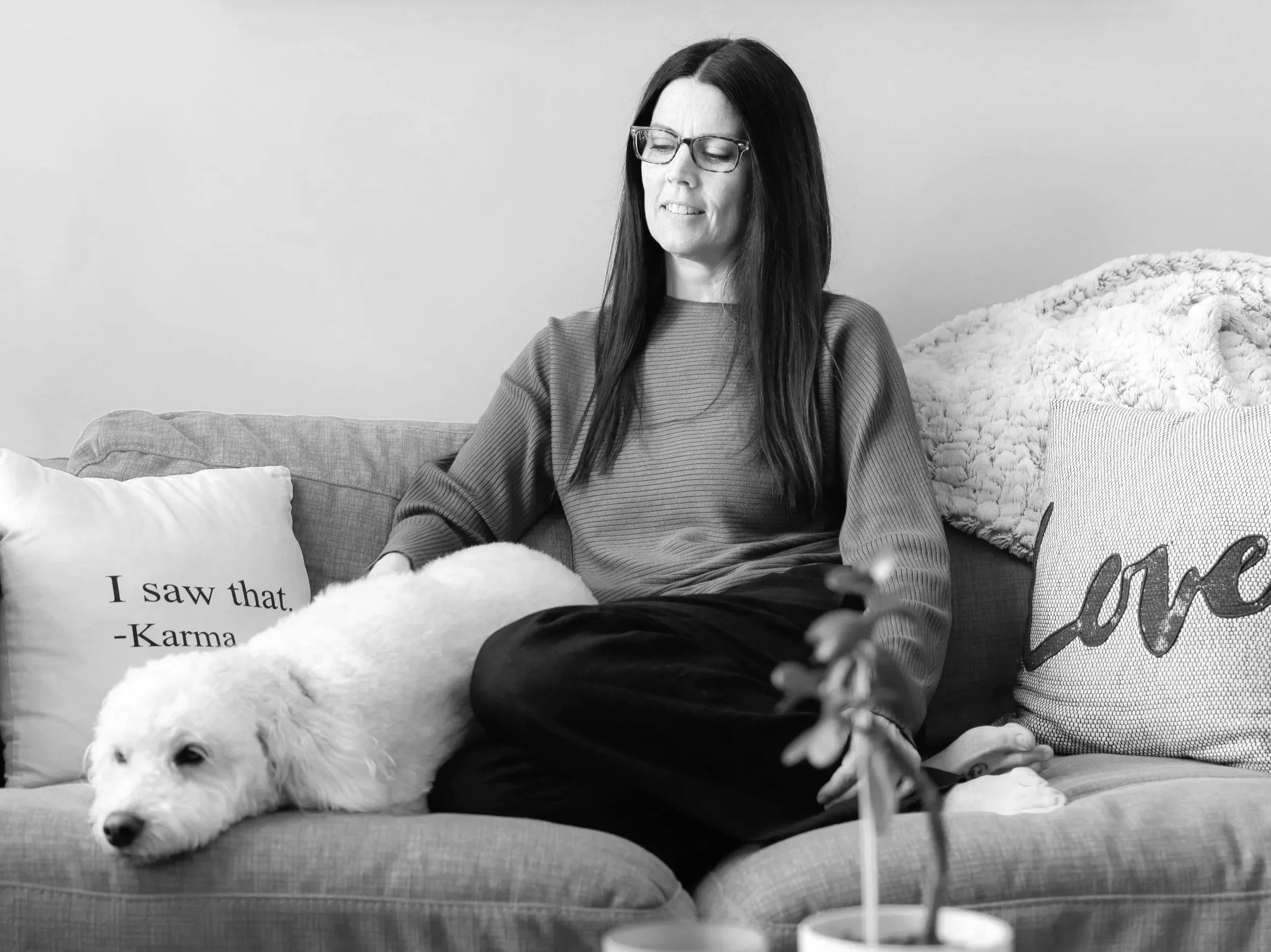 A woman with long dark hair, glasses, and a striped shirt sitting on a couch with a white dog lying beside her. There are decorative pillows behind her, one with the text "I saw that. -Karma" and another with the word "Love." The woman appears to be gazing at a smartphone in her hand.