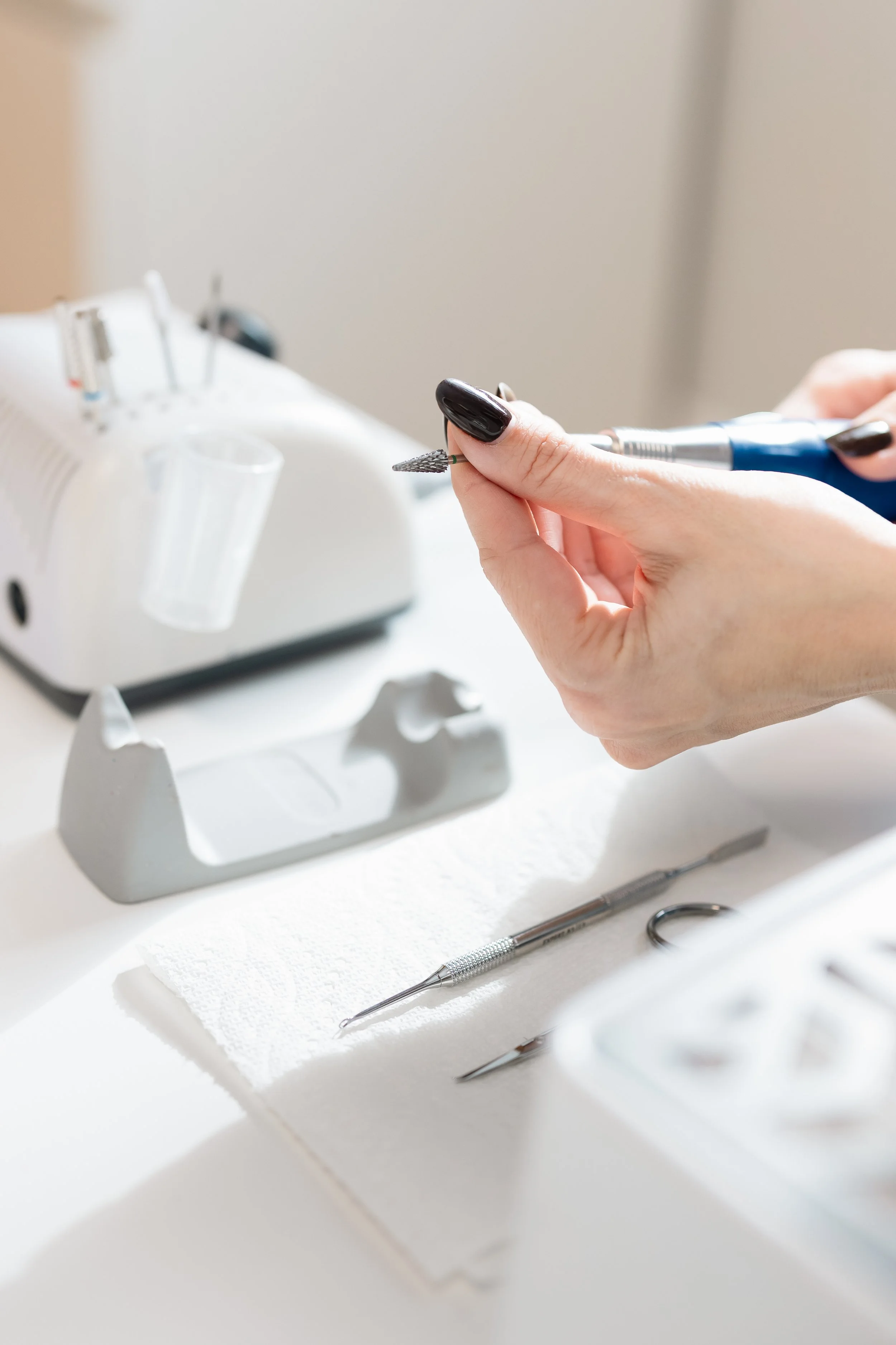 Close-up of a person's hands holding a manicure drill, with manicure tools and a UV lamp on a white table.
