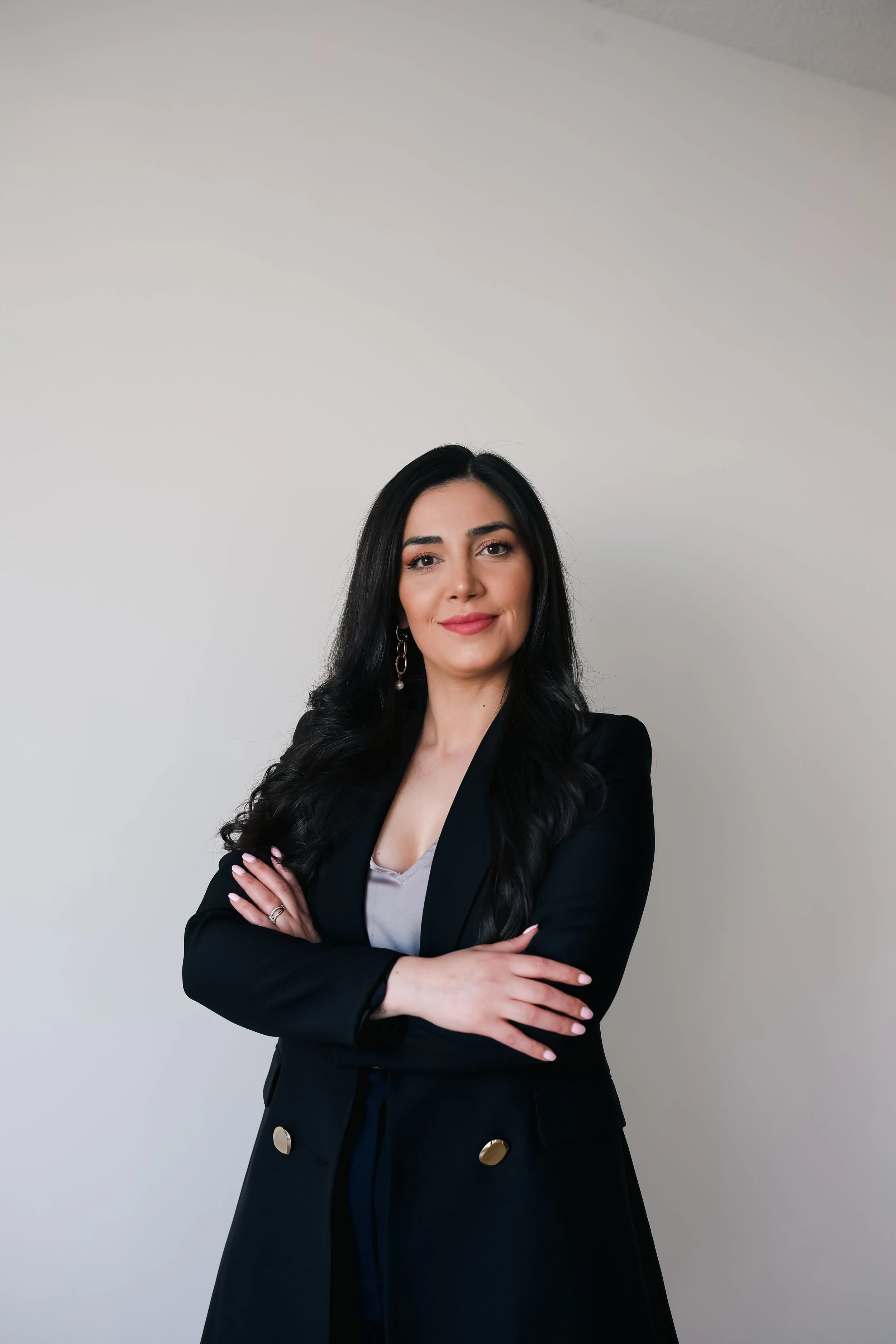 A woman with long black hair, wearing a black blazer with gold buttons, poses with crossed arms against a plain white wall.