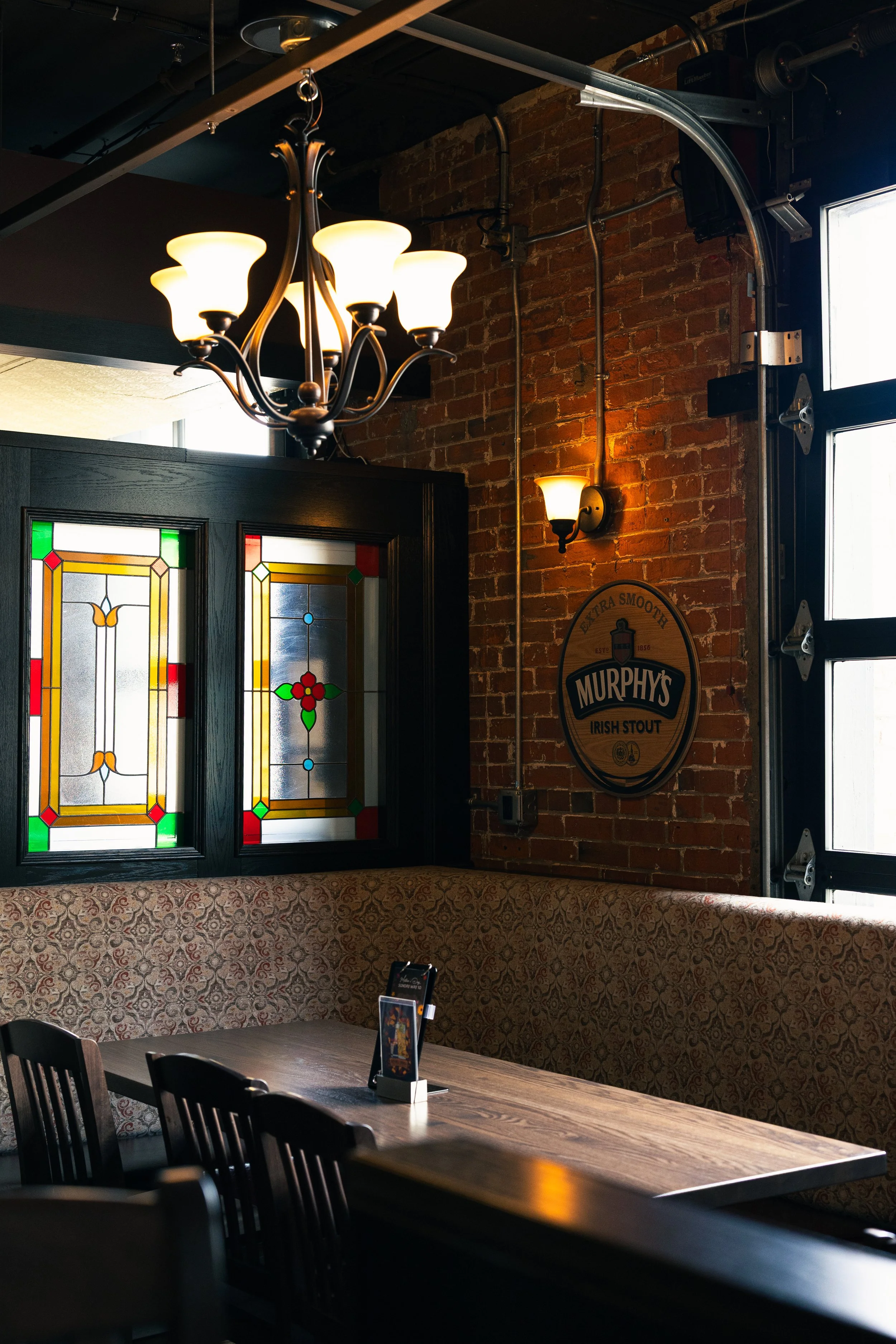 Interior of a Irish pub with a lit chandelier, stained glass windows, a brick wall, wooden tables and chairs, and a Murphy's Irish Stout sign on the wall.