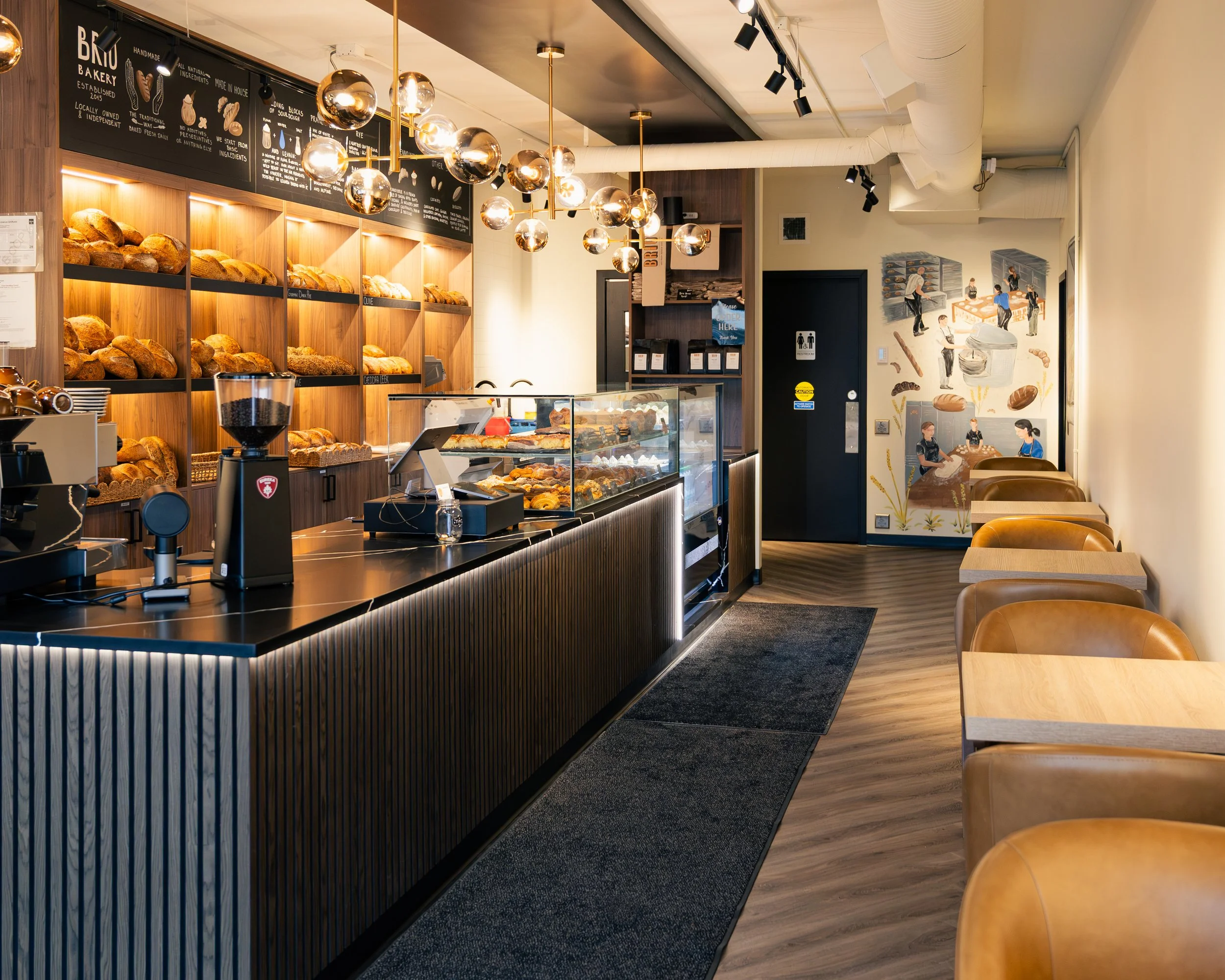 Interior of a bakery café with baked bread on wooden shelves, modern hanging light fixtures, a black counter with baked goods in display cases, and seating area with light-colored chairs and tables. Wall art depicts bakers and baked goods.