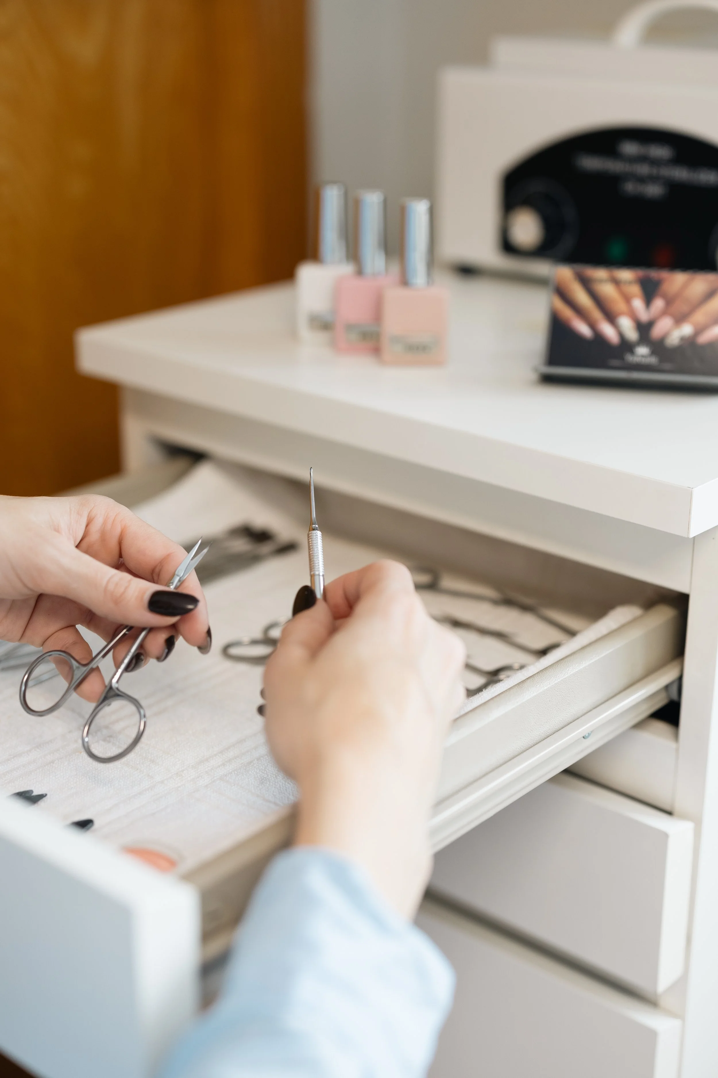 A person’s hands holding nail cuticle scissors and a cuticle pusher over a white drawer filled with nail art supplies.