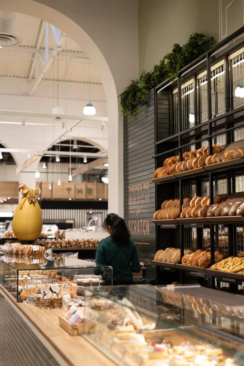 A woman in a dark green sweater is shopping in a bakery with shelves filled with loaves of bread, pastries, and baked goods, inside a modern bakery or cafe.