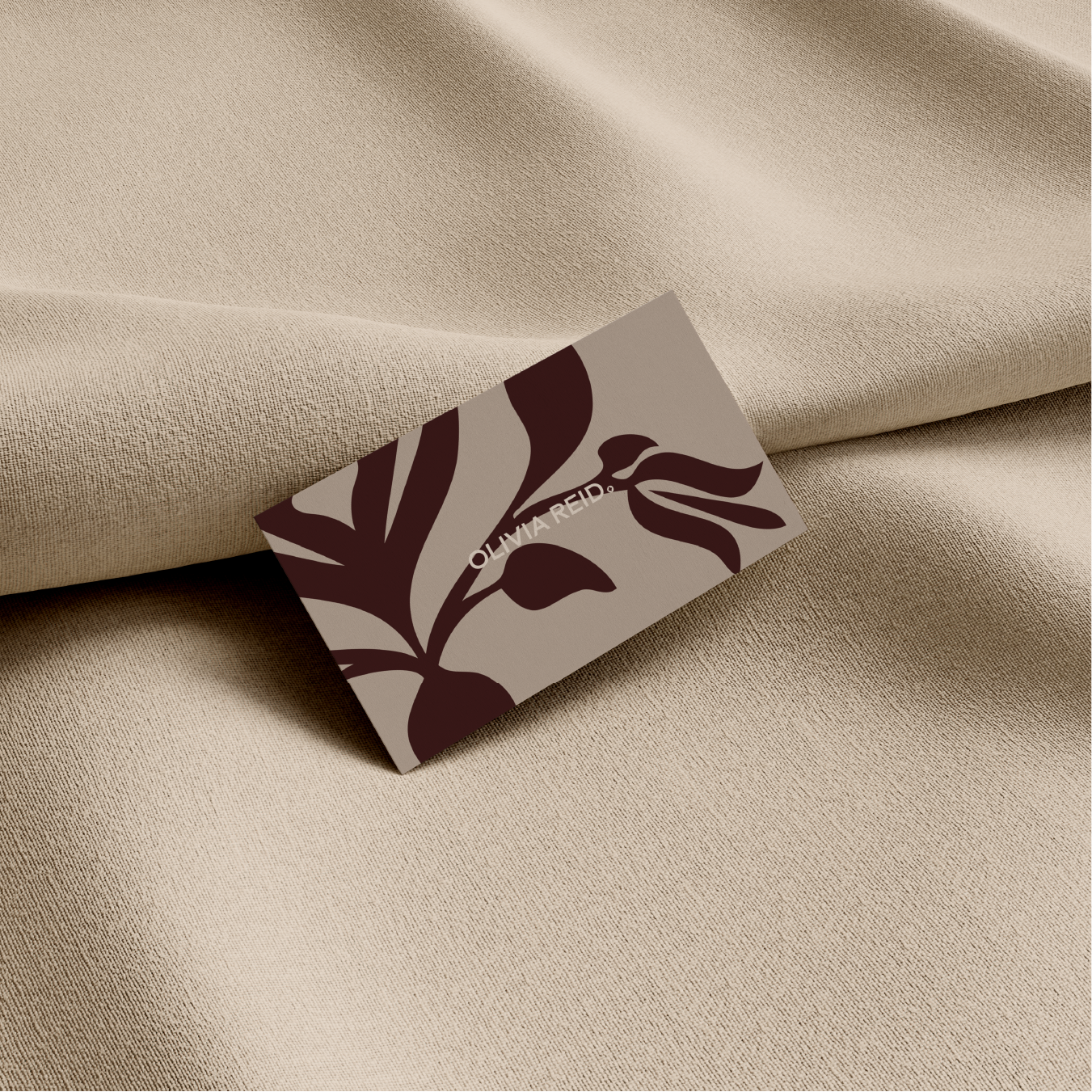 A beige textured fabric with a sign that has a brown and beige leaf pattern and the text 'Olivia Reid' on it, placed on the fabric.