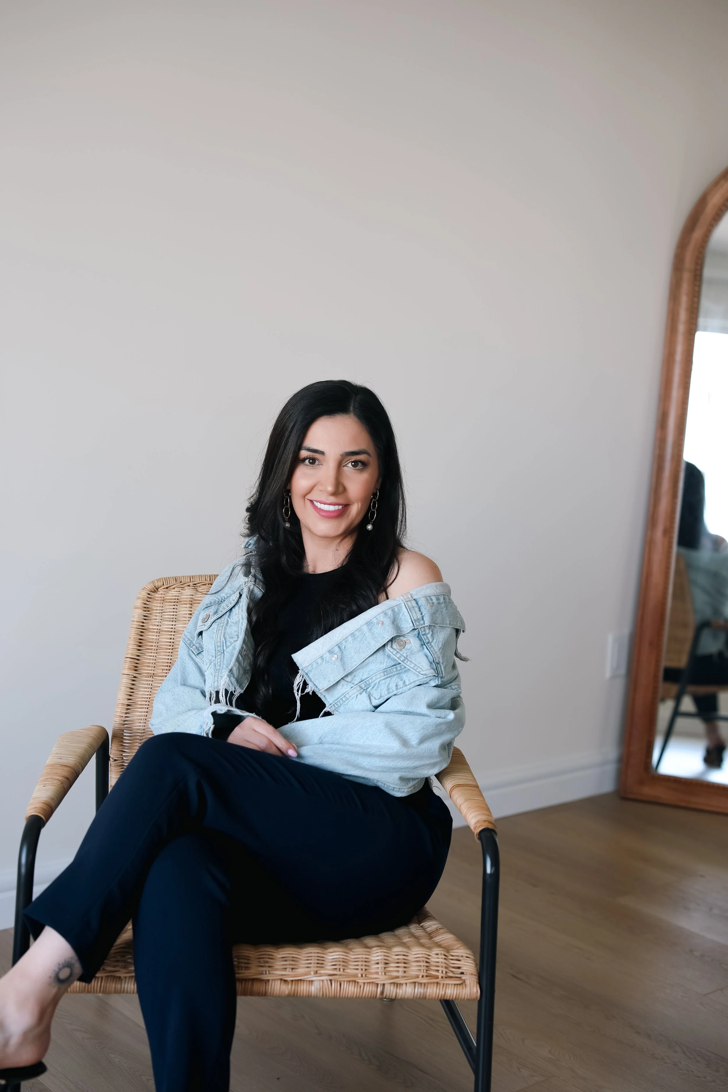 A woman sitting on a woven chair in front of a plain wall, smiling and looking at the camera.