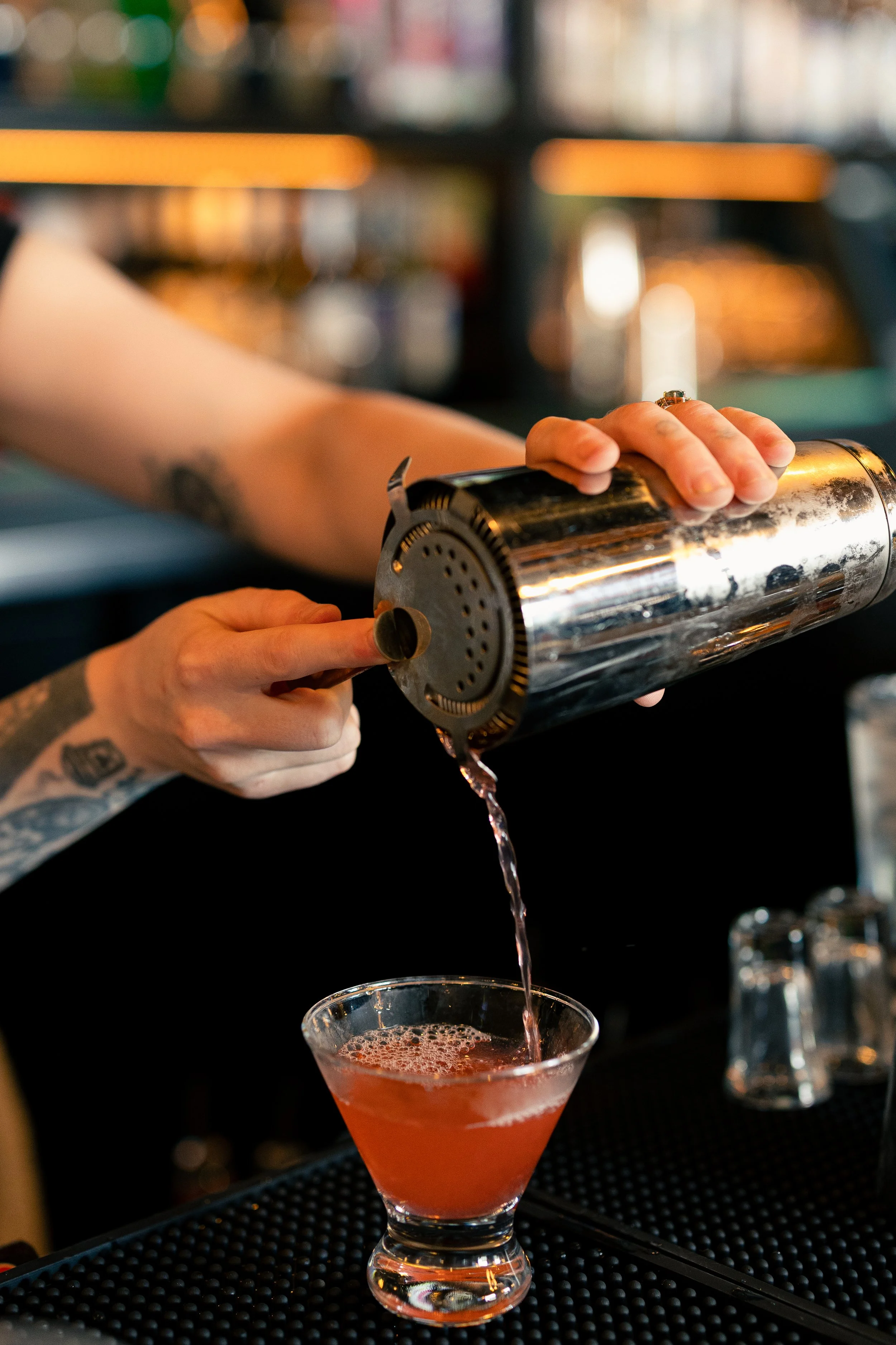 A bartender pours a pink cocktail from a metal cocktail shaker into a martini glass, with a blurred bar background.