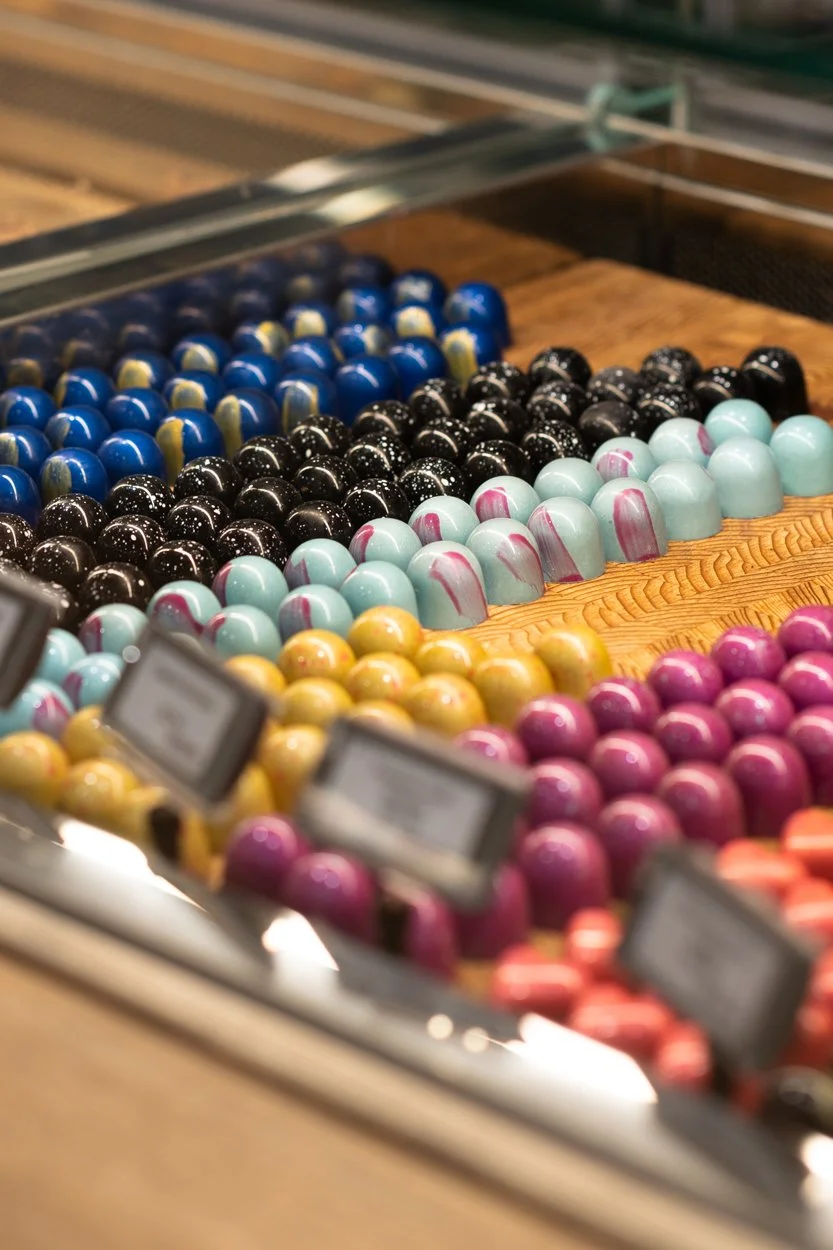 A display of colorful round chocolates in various colors including blue, black, light blue, yellow, pink, and purple, arranged in rows at a candy store.