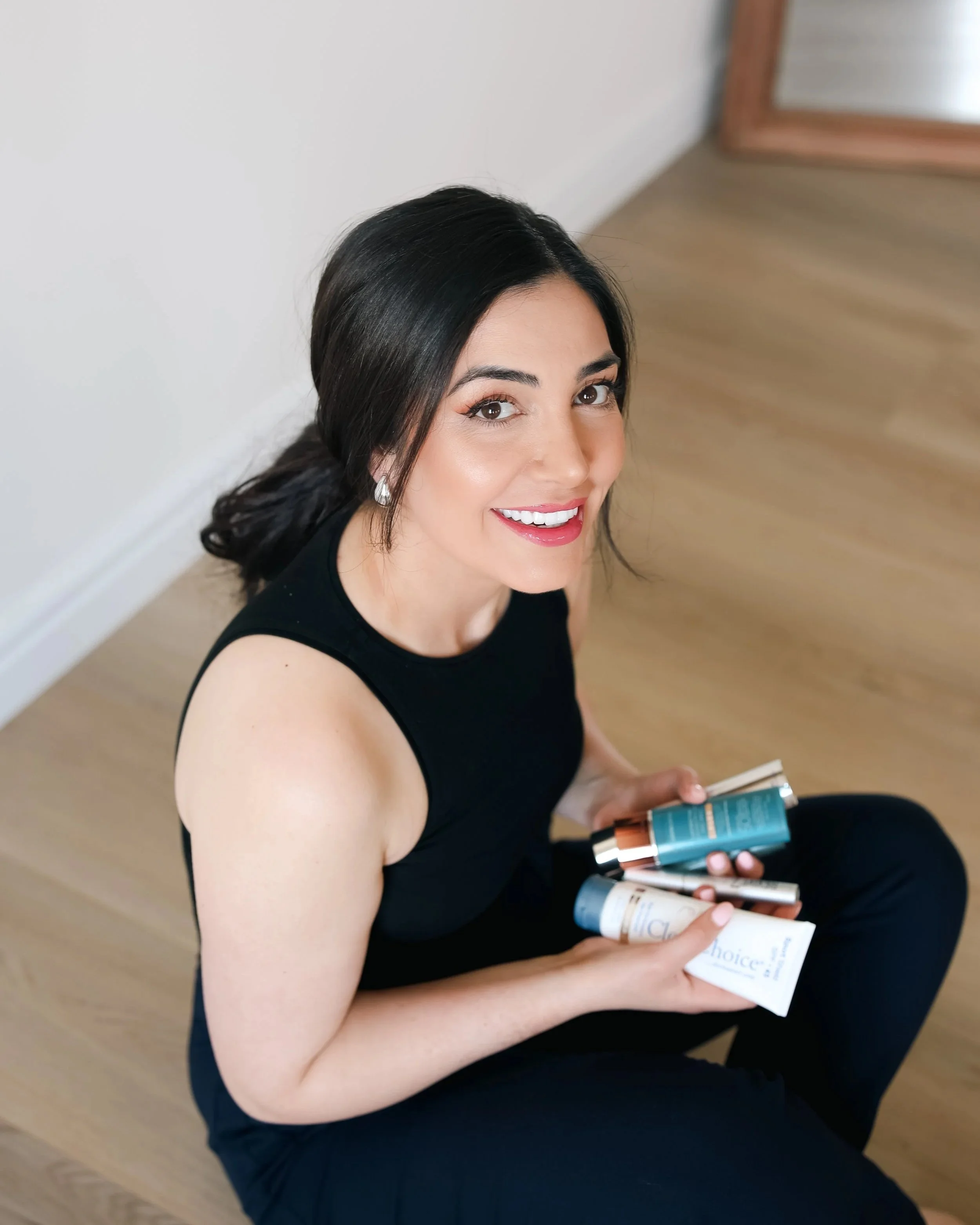A smiling woman with dark hair, wearing a black sleeveless top, sitting on a wooden floor, holding several skincare or cosmetic products in her hands.