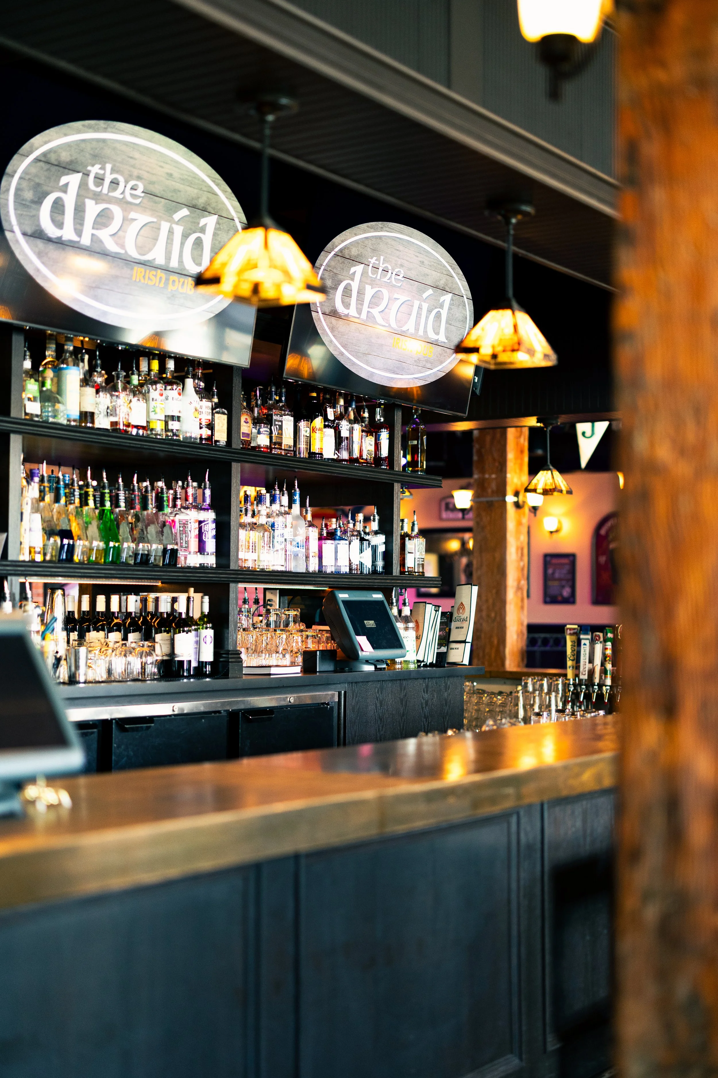 Interior view of a bar at 'The Druid,' an Irish pub, showing shelves with liquor bottles, a cash register, and ambient lighting with hanging lamps.