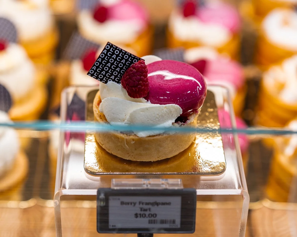 Close-up of a berry framboise tart with raspberry, white cream, and glossy pink glaze on top, displayed on a transparent stand in a bakery.