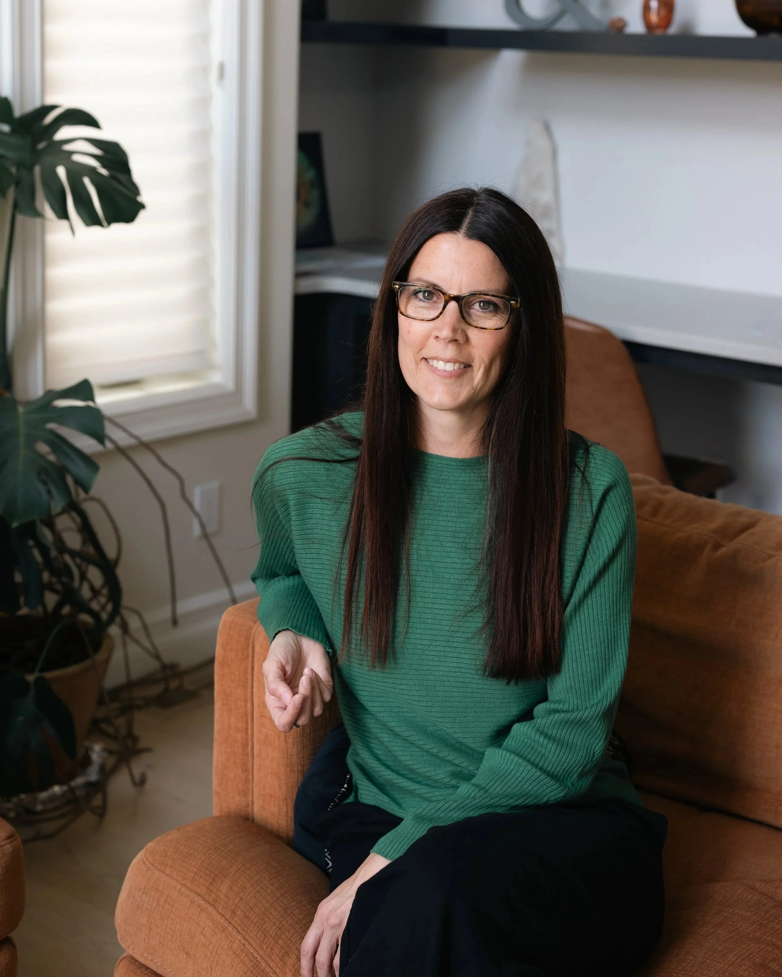 A woman with long dark hair and glasses sitting on an orange sofa in a room with a window and houseplants.
