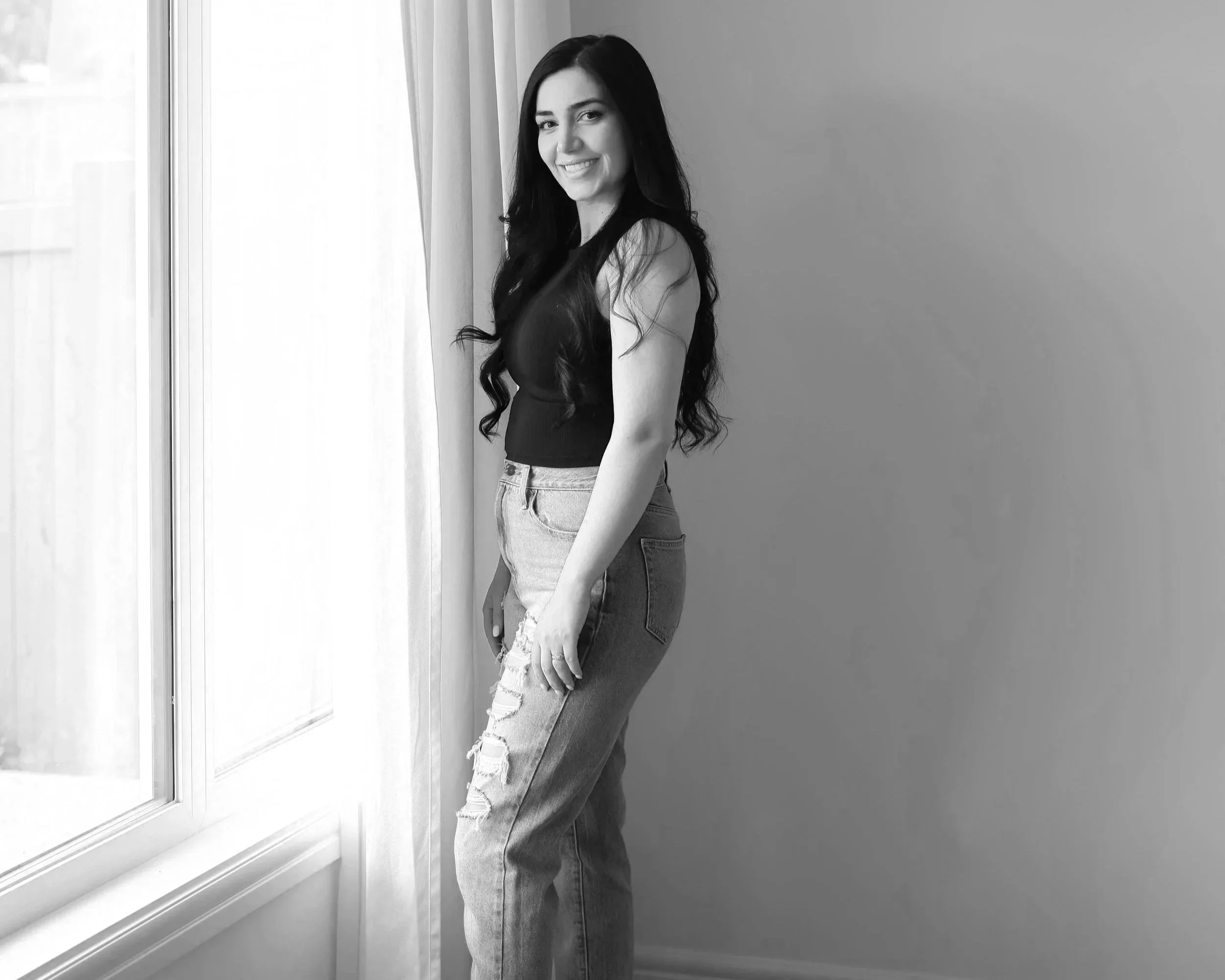 A smiling woman with long dark hair, wearing a sleeveless top and ripped jeans, standing beside a window in a room with plain walls.
