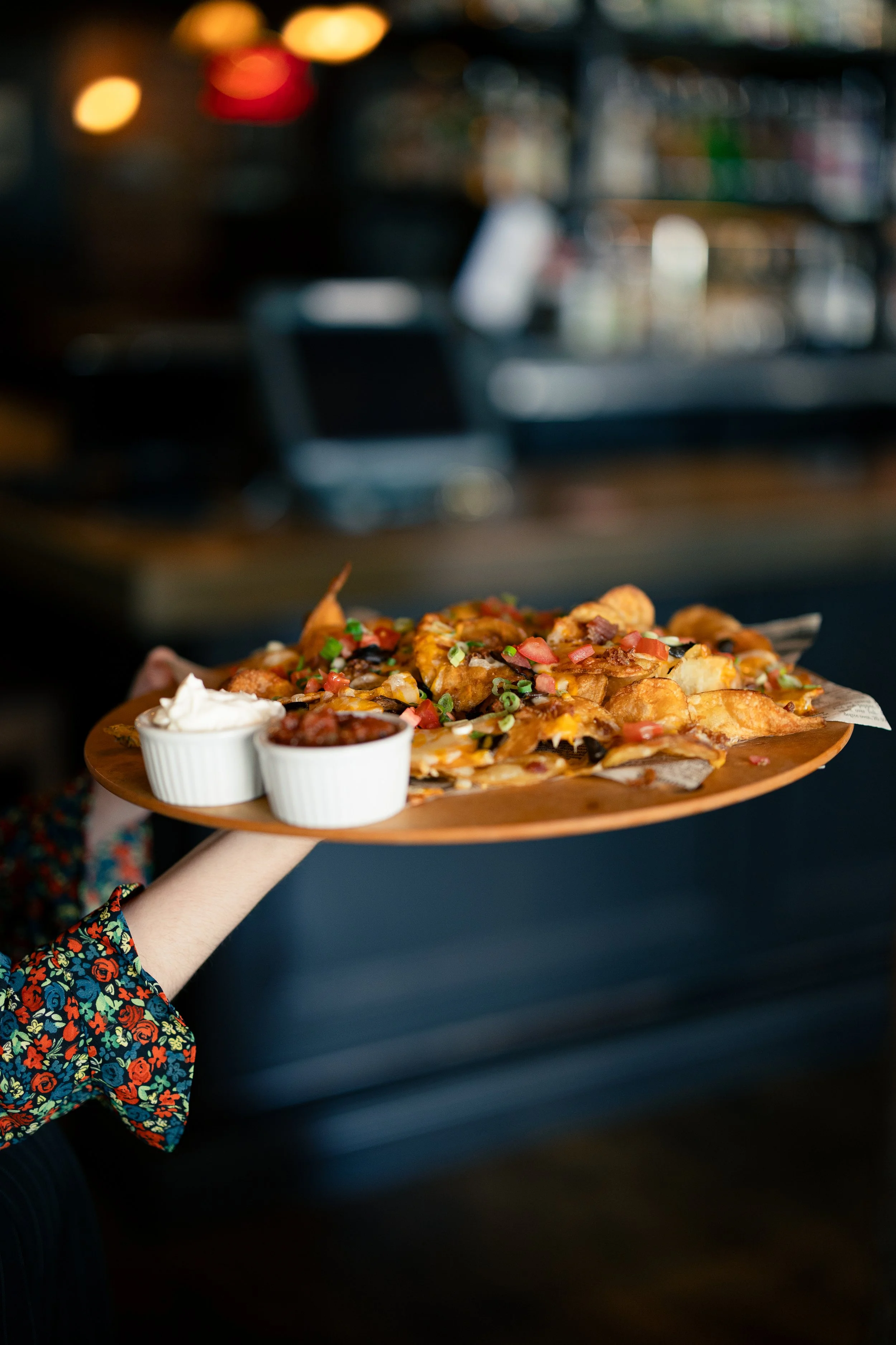 A person wearing a colorful floral sleeve is holding a wooden tray with nachos, topped with diced tomatoes, green onions, cheese, and served with sour cream and salsa, in a dimly lit restaurant or bar.