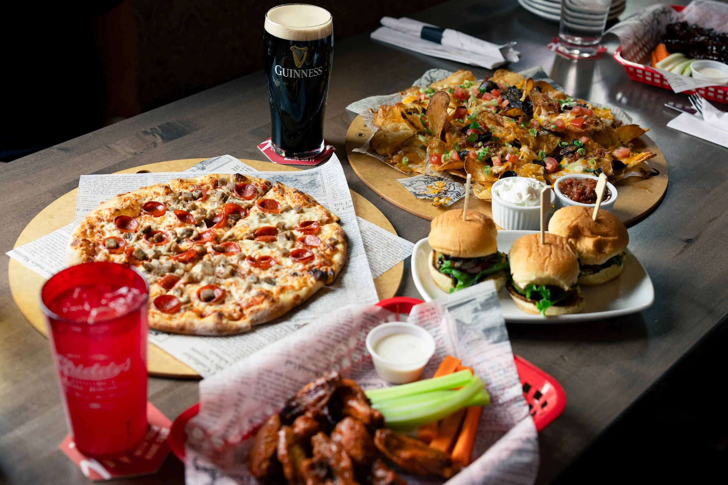 Various dishes on a table including a pepperoni pizza, nachos topped with cheese and vegetables, mini sliders, a glass of Guinness beer, a glass of cola, dipping sauces, and celery sticks.