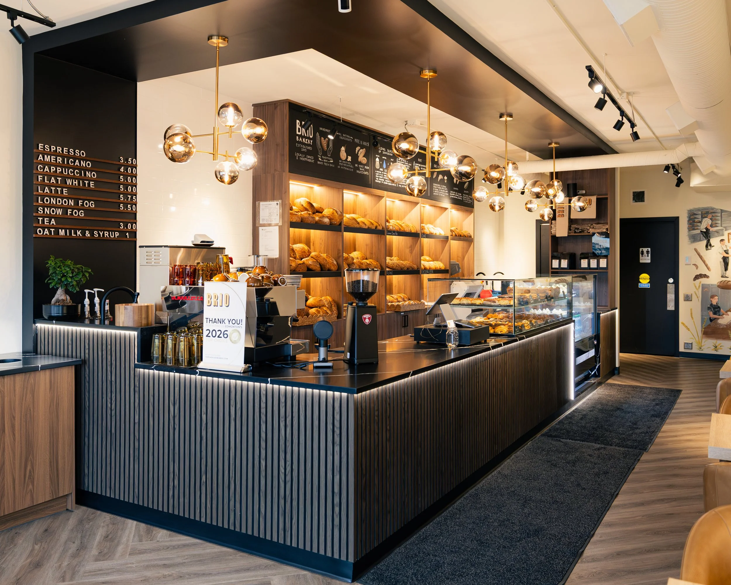Interior of a modern bakery with a black and wooden display counter, illuminated by multiple gold pendant lights, showcasing baked bread and pastries. A black espresso menu board is on the wall, and there are small decorative plants and a sign on the counter.