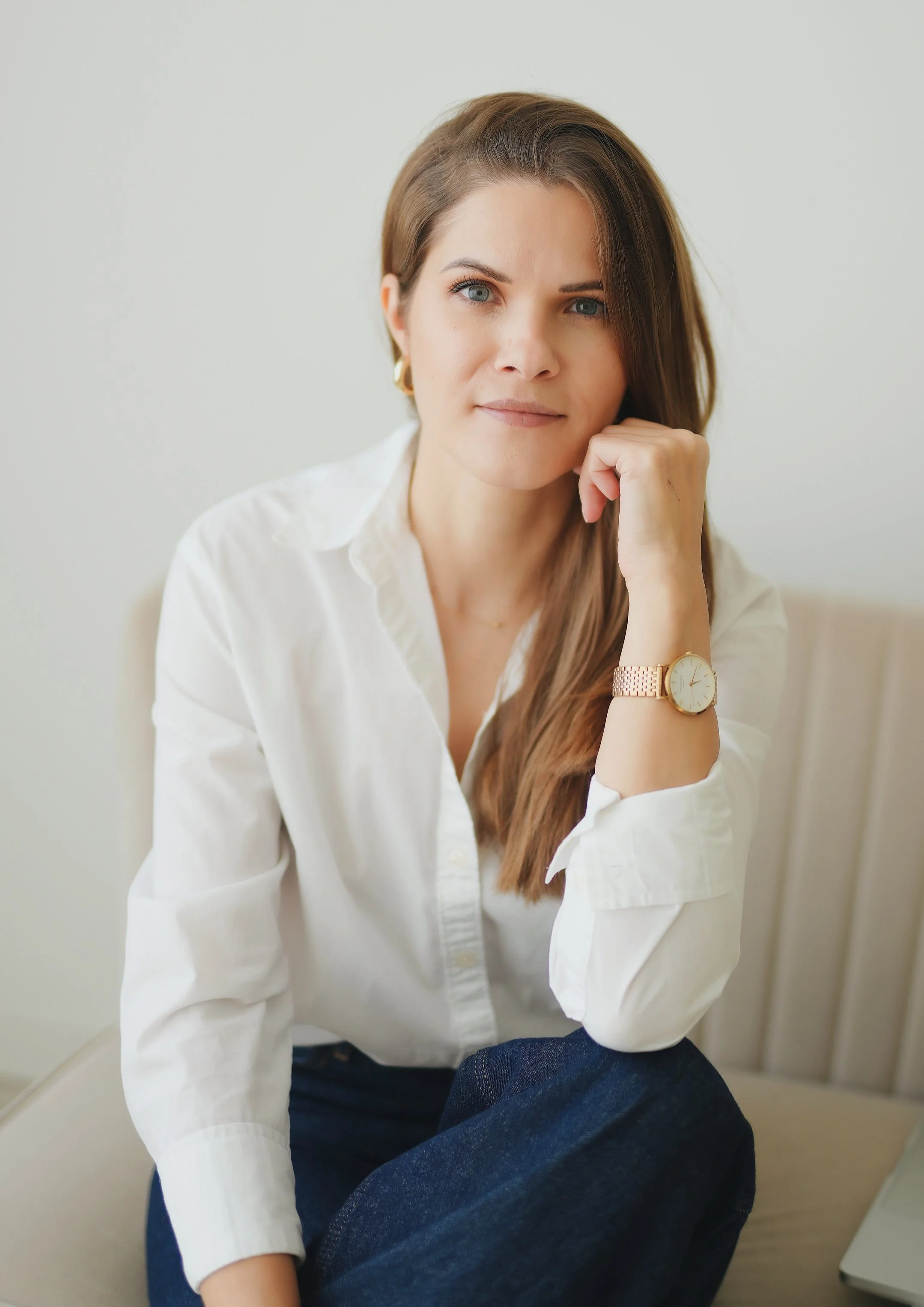 A woman with long brown hair and blue eyes, wearing a white shirt, a gold watch, and gold earrings, sitting on a beige chair with her hand resting under her chin, looking directly at the camera.