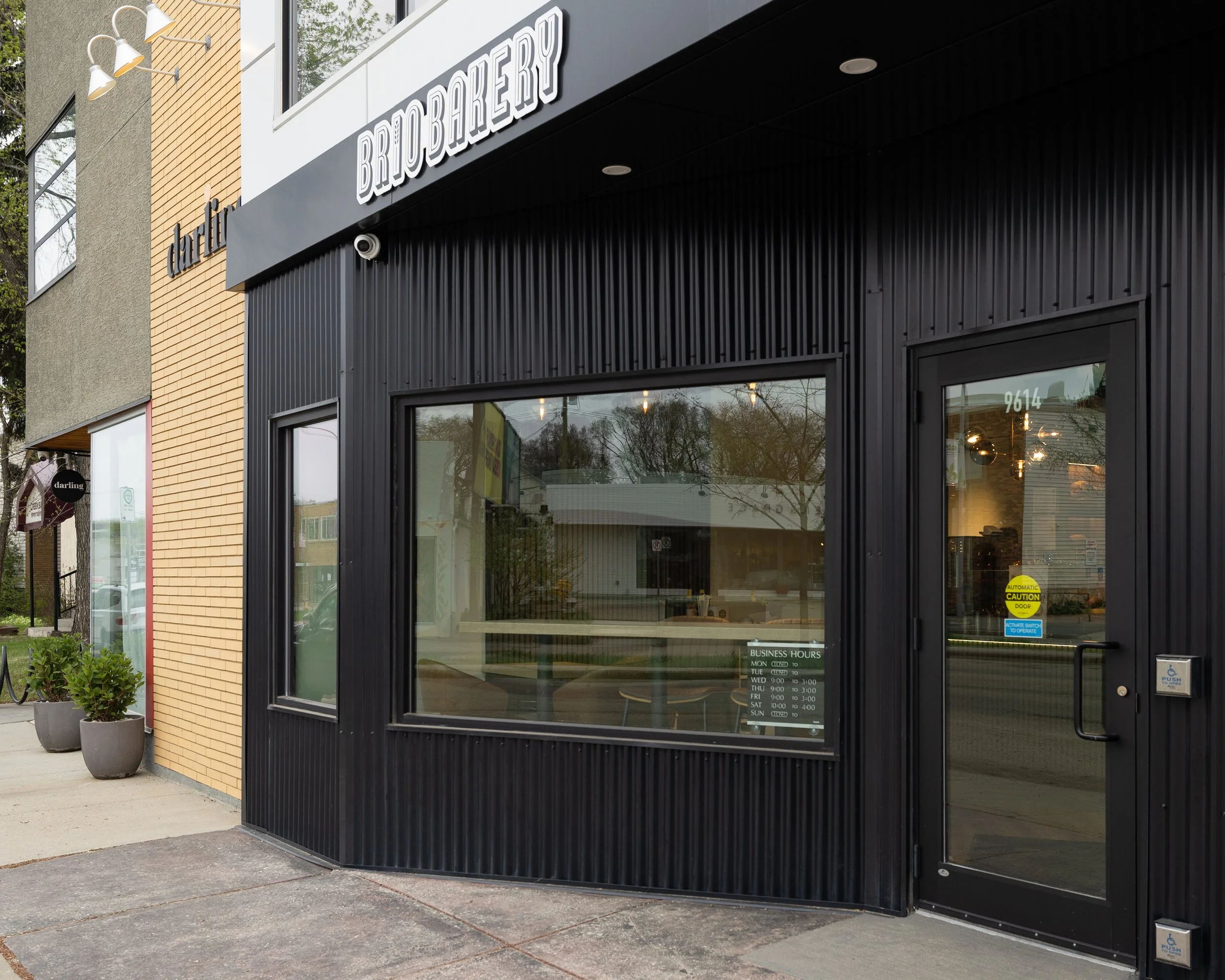 Storefront with black exterior and large windows, sign reading 'BRIO BAKERY,' potted plants outside, business hours sign, and reflections in the windows.