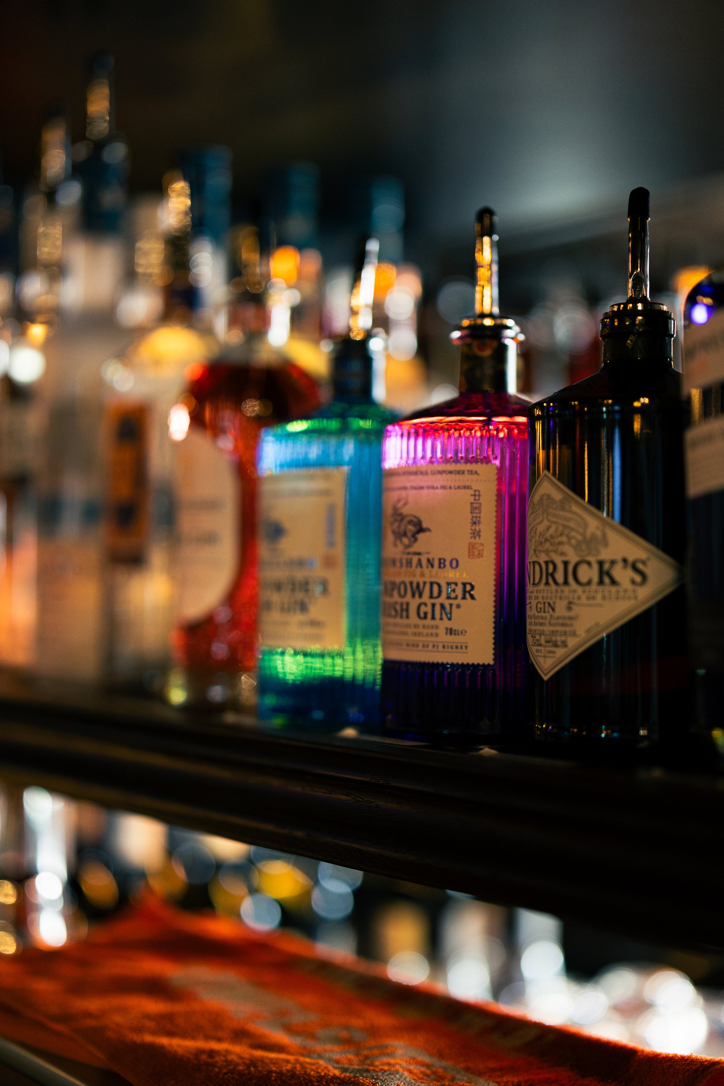 Blurred shelves of colorful bottles of alcohol, including gin, on a bar or liquor store shelf, with a dark background and bokeh light effects.