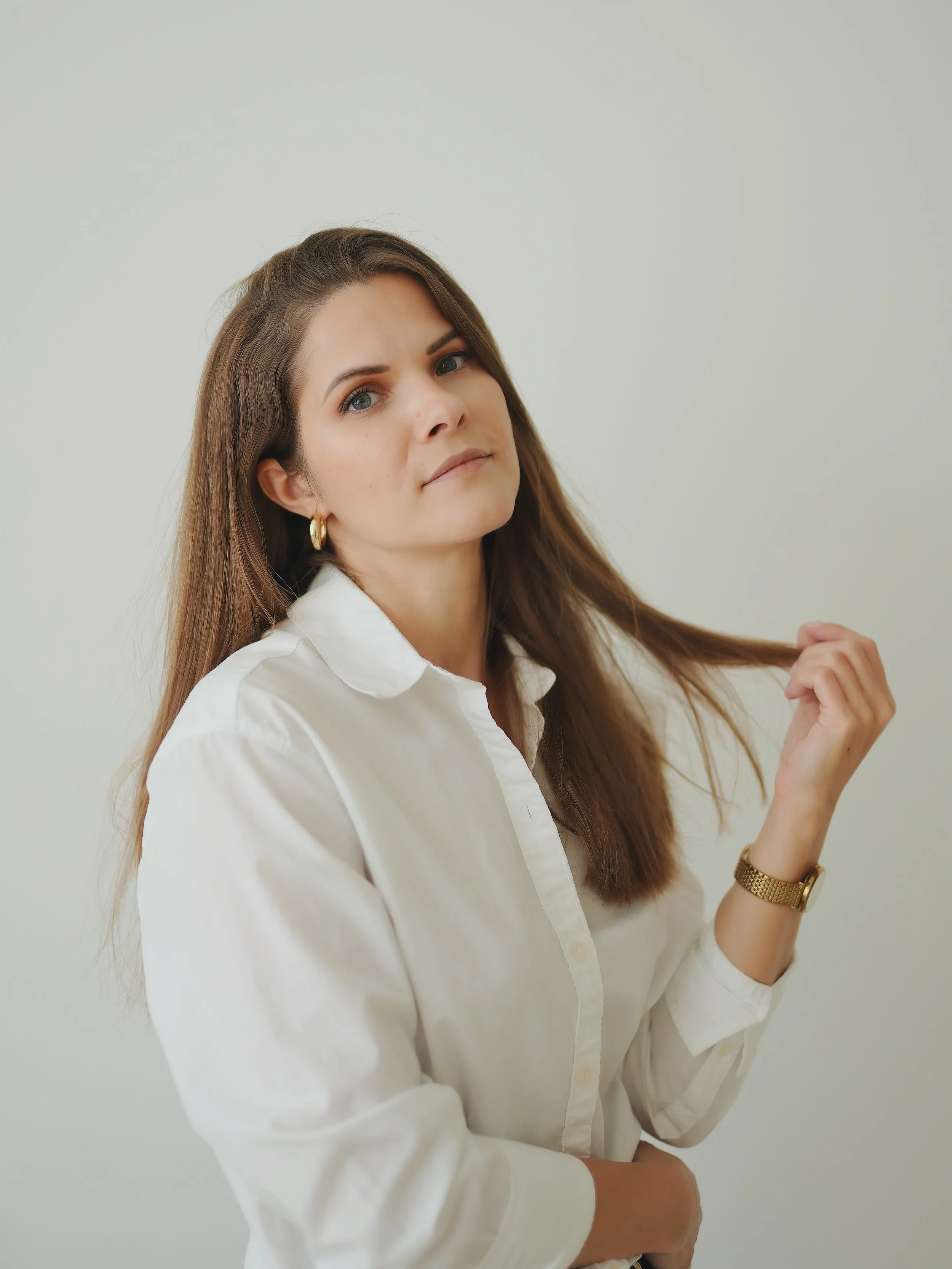 A woman with long brown hair, blue eyes, wearing a white button-up shirt, gold hoop earrings, and a gold watch, standing against a plain light-colored background, holding a strand of her hair.