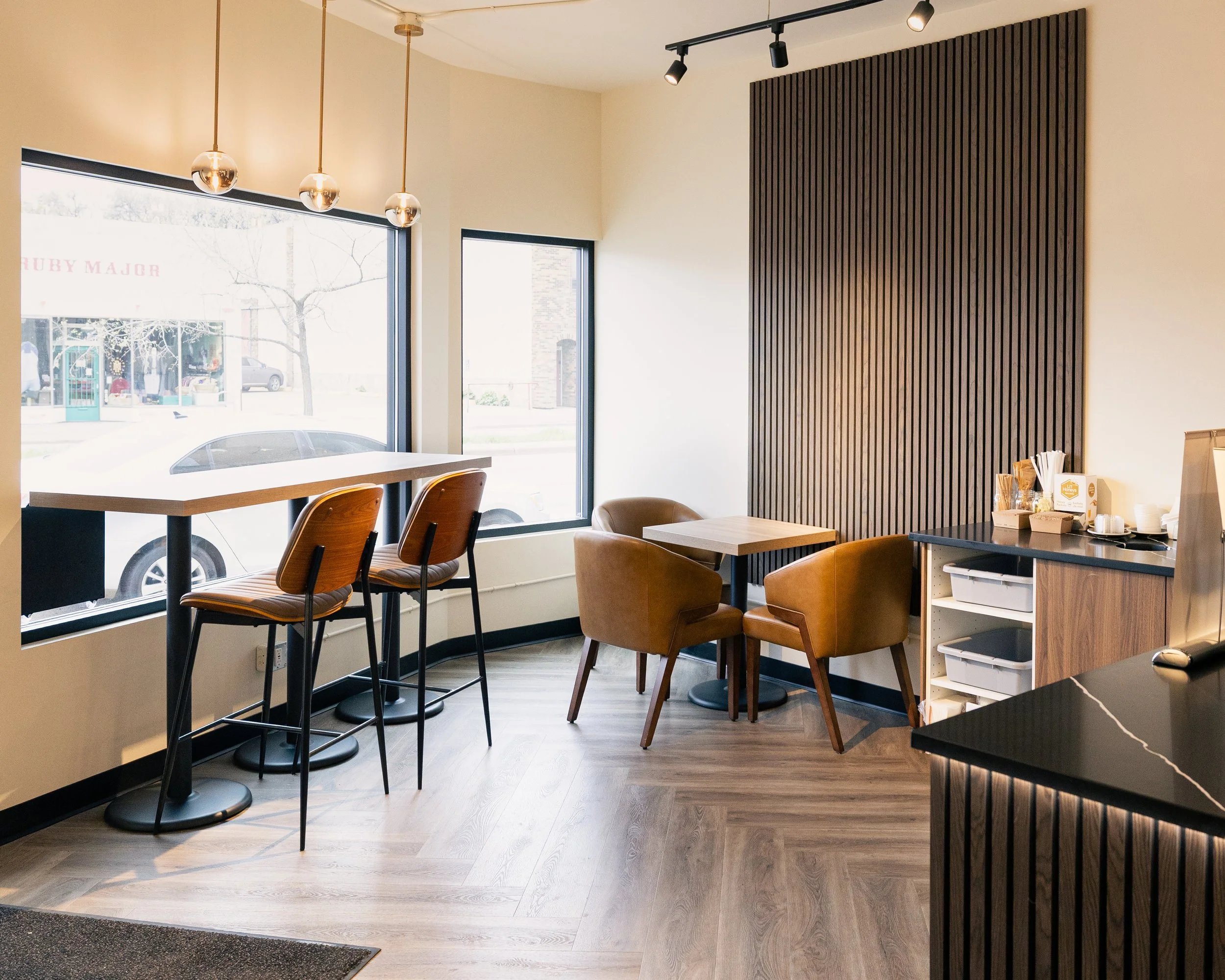Interior of a modern cafe with large windows, wooden tables, and chairs, with a dark wood accent wall and track lighting.